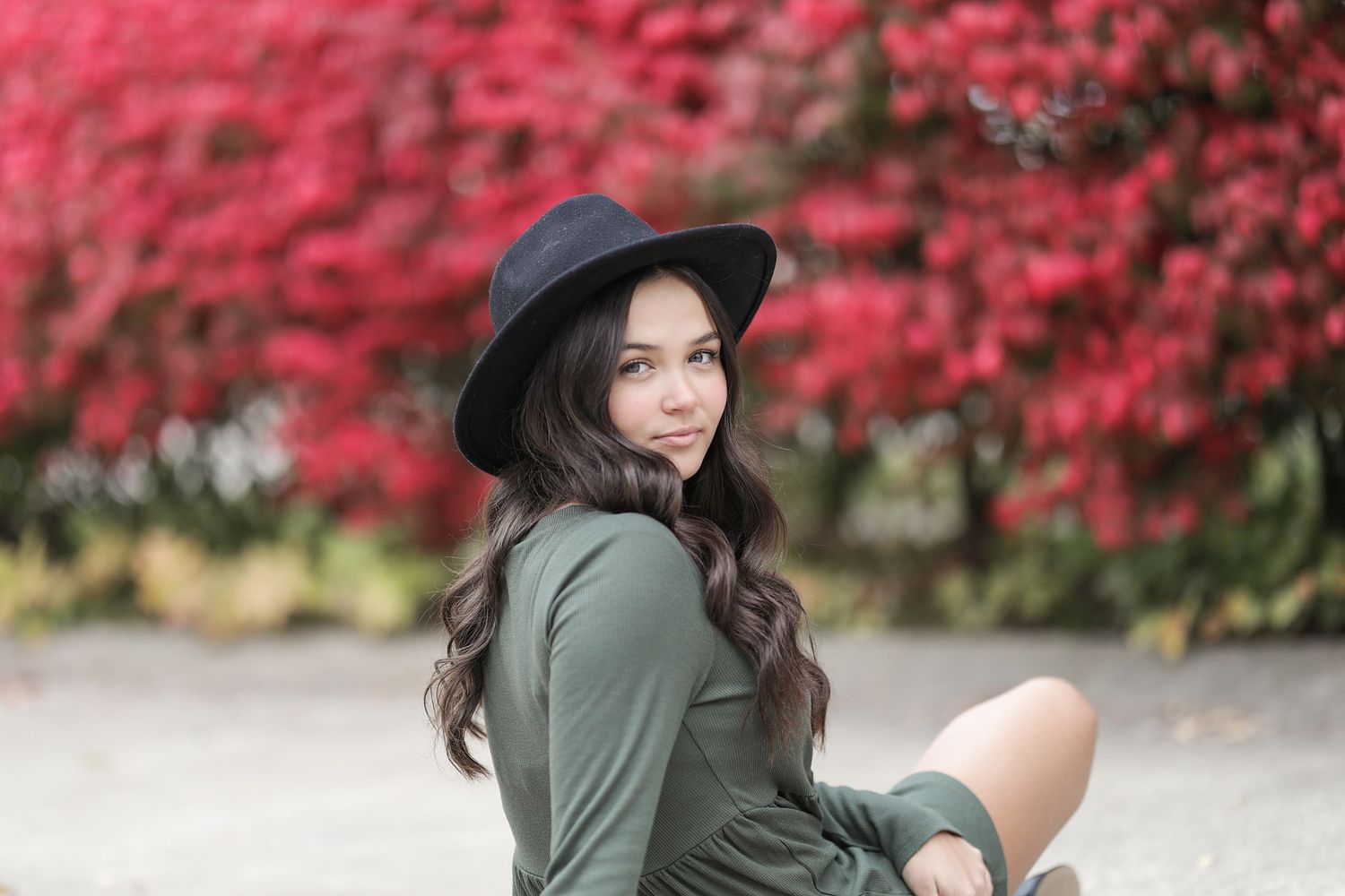 A person in a hat sits outdoors against a backdrop of vibrant red autumn foliage.