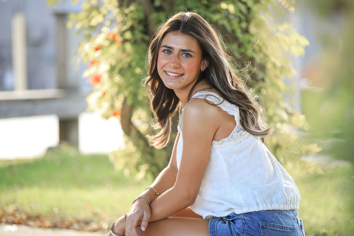 Woman wearing white top and denim shorts smiles while sitting outdoors in natural sunlit setting.