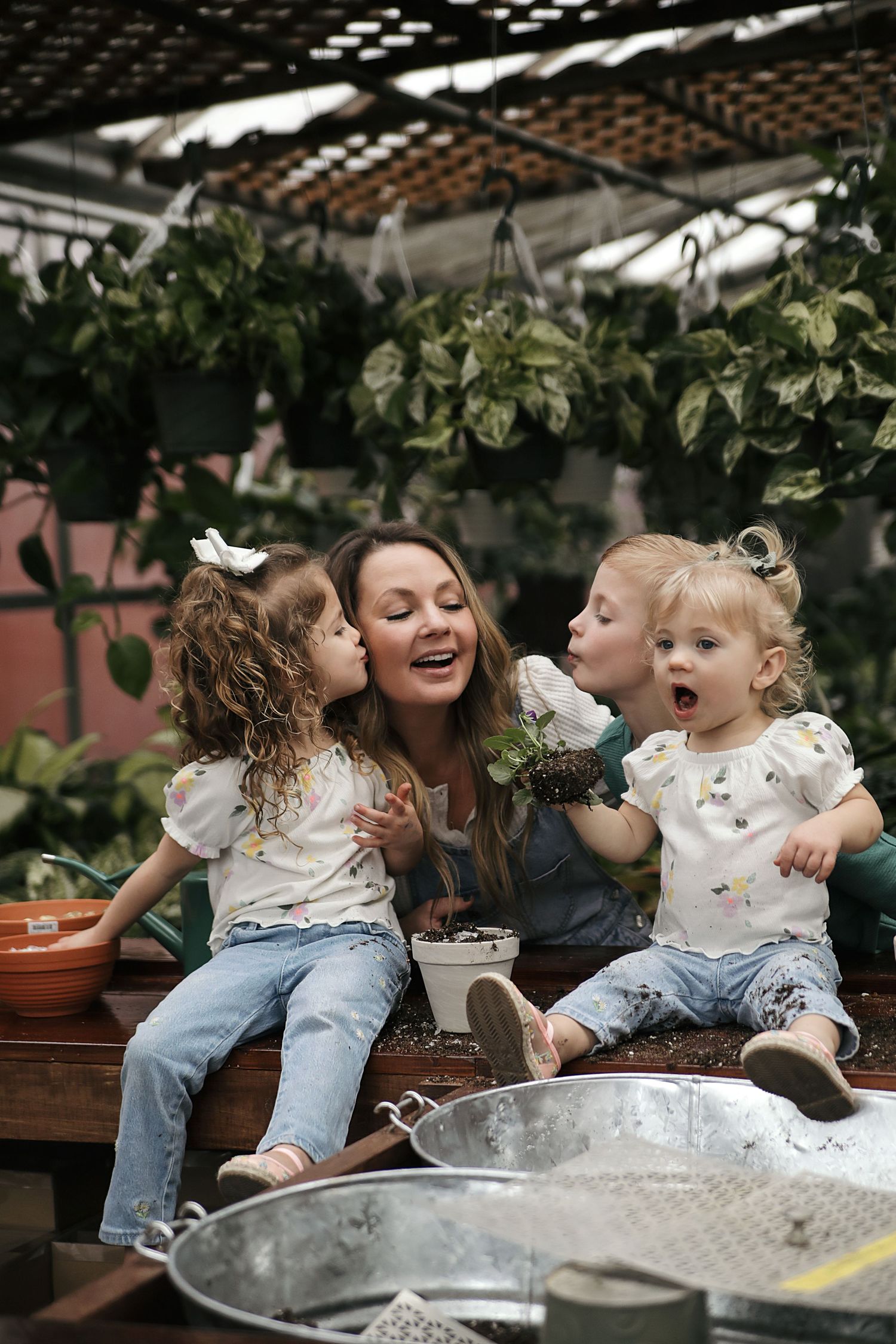Three people share joyful moments together while sitting outdoors among potted plants and hanging greenery.