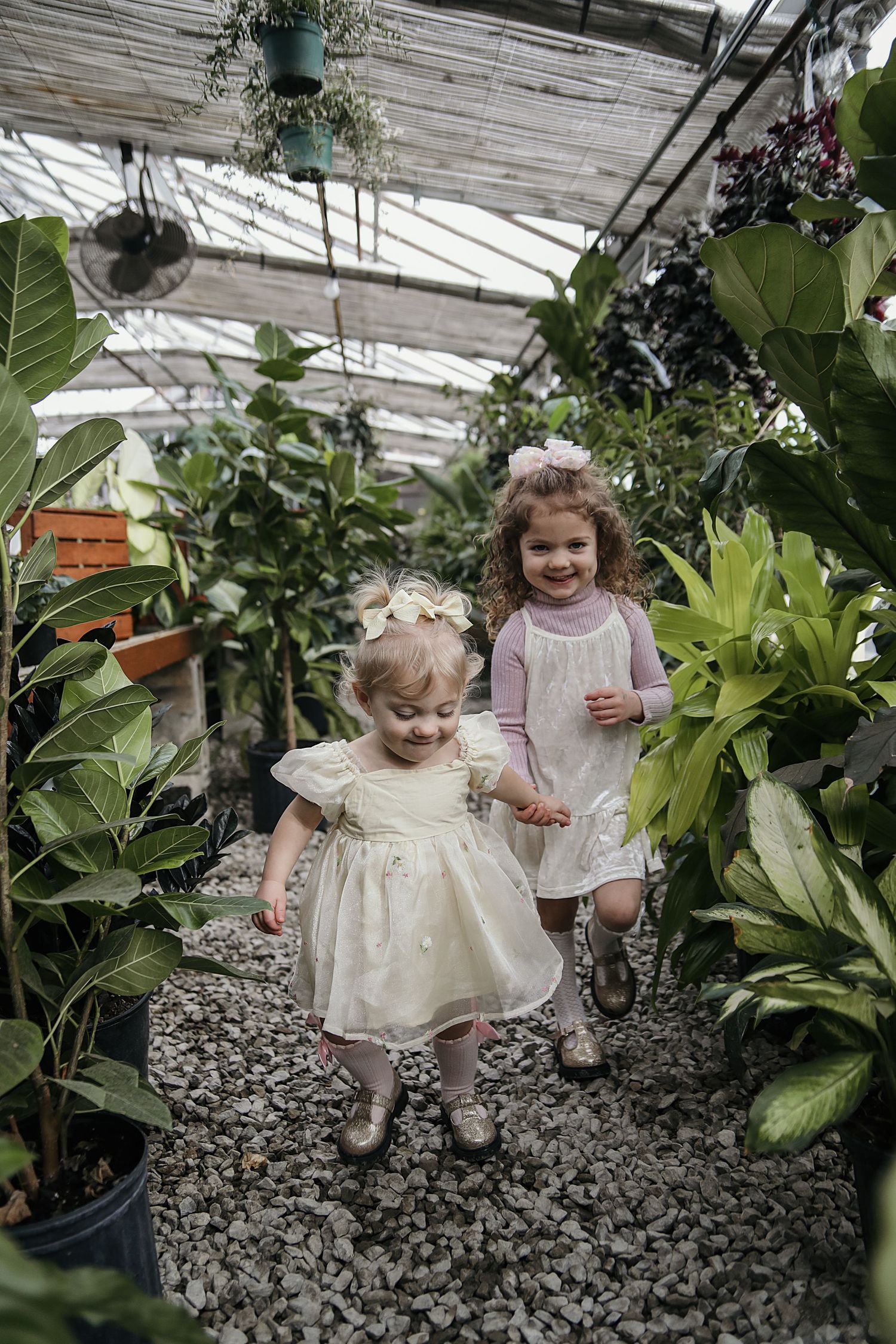 Two small children explore a greenhouse filled with lush plants and greenery on a gravel path.