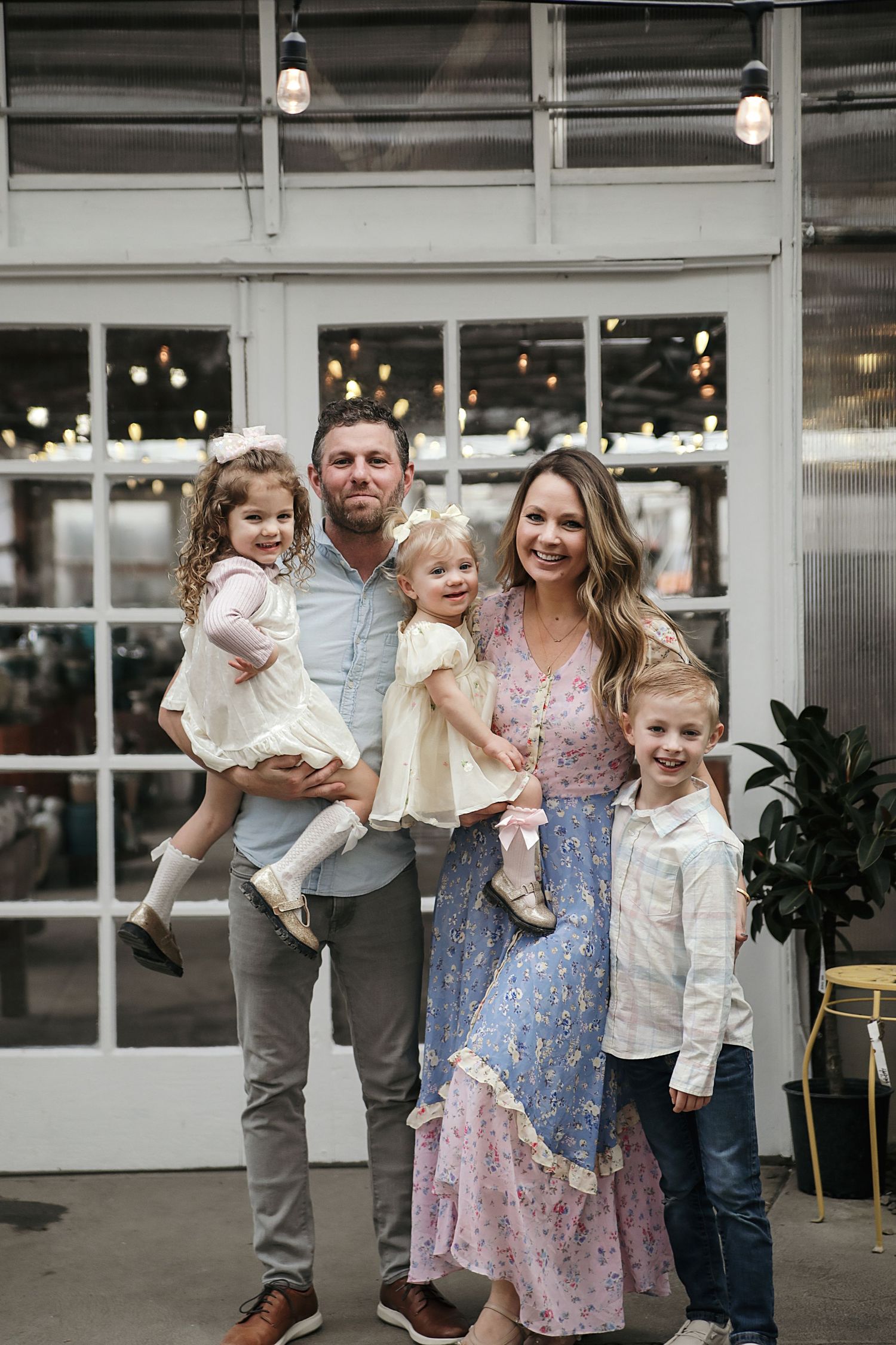 A family of five poses together in front of a glass storefront with twinkling lights in the background.