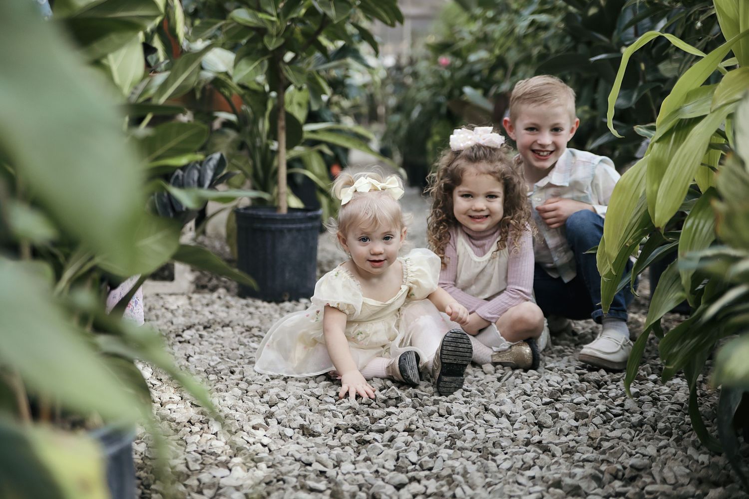 Three young children sitting and playing together on gravel path surrounded by green plants and foliage.