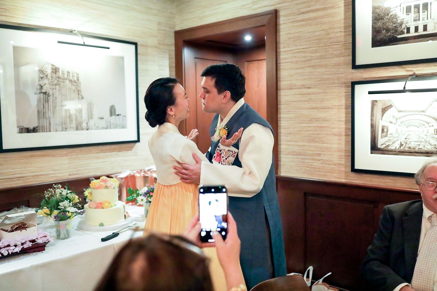 Couple shares an intimate moment at a restaurant while someone takes a photo with their phone.