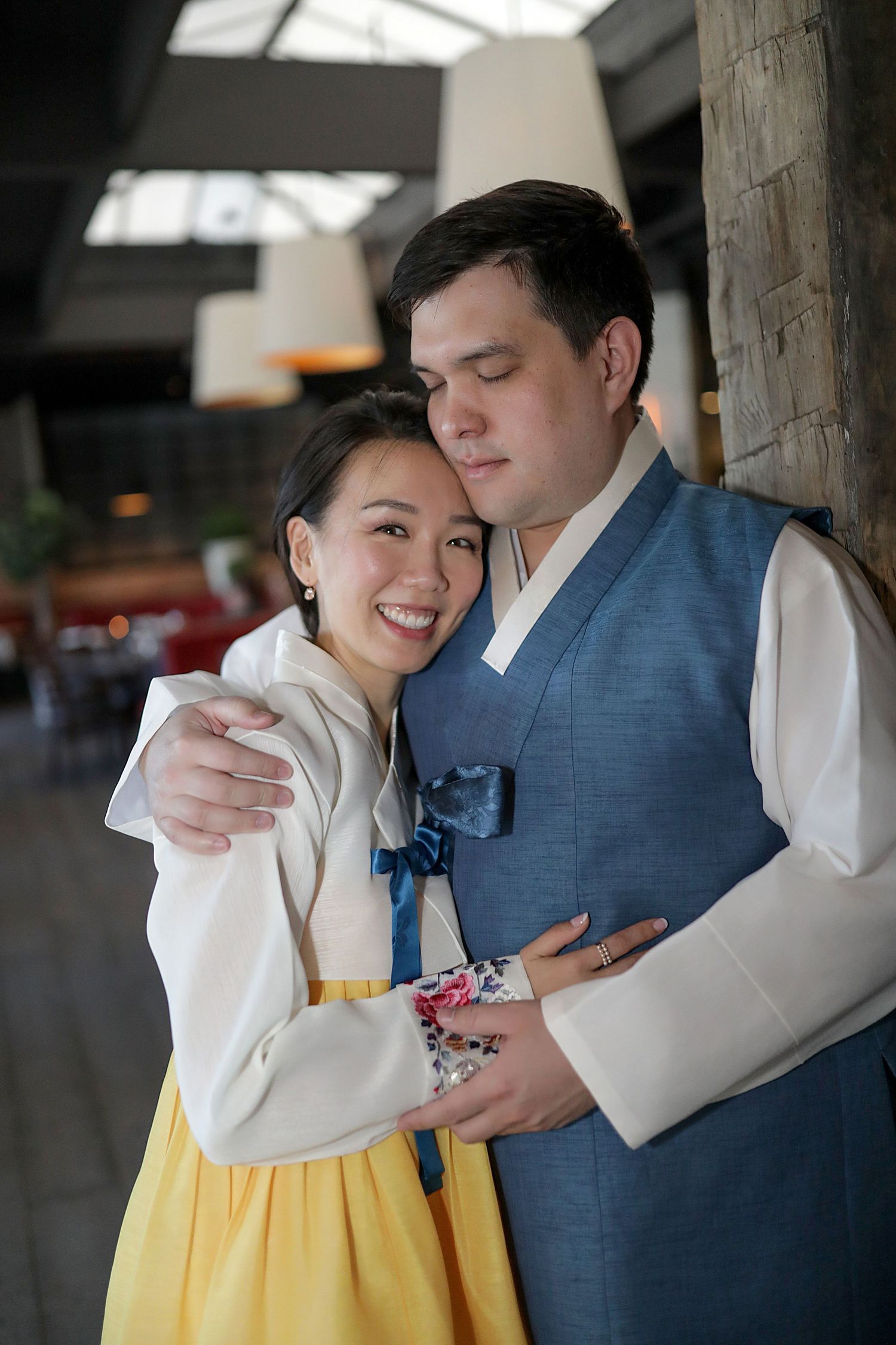 A couple in traditional Korean hanbok embrace while wearing contrasting blue and yellow formal attire.