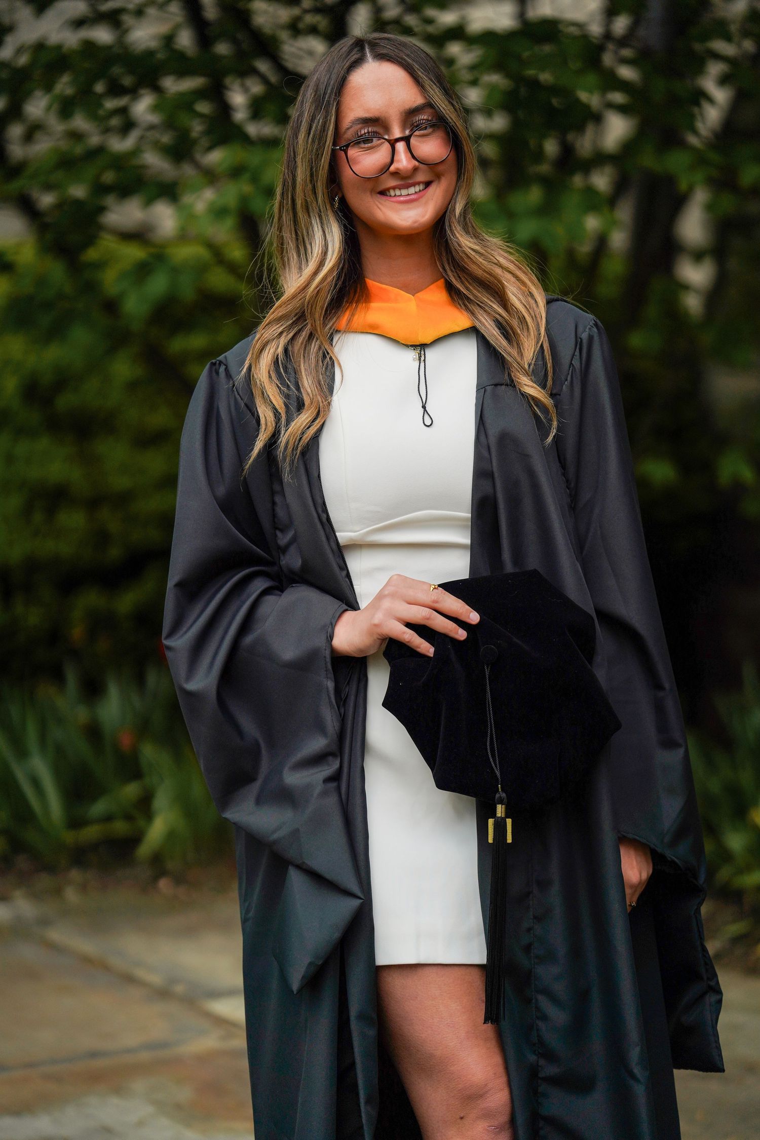 Graduate in black academic regalia holds white diploma holder in outdoor setting surrounded by greenery.