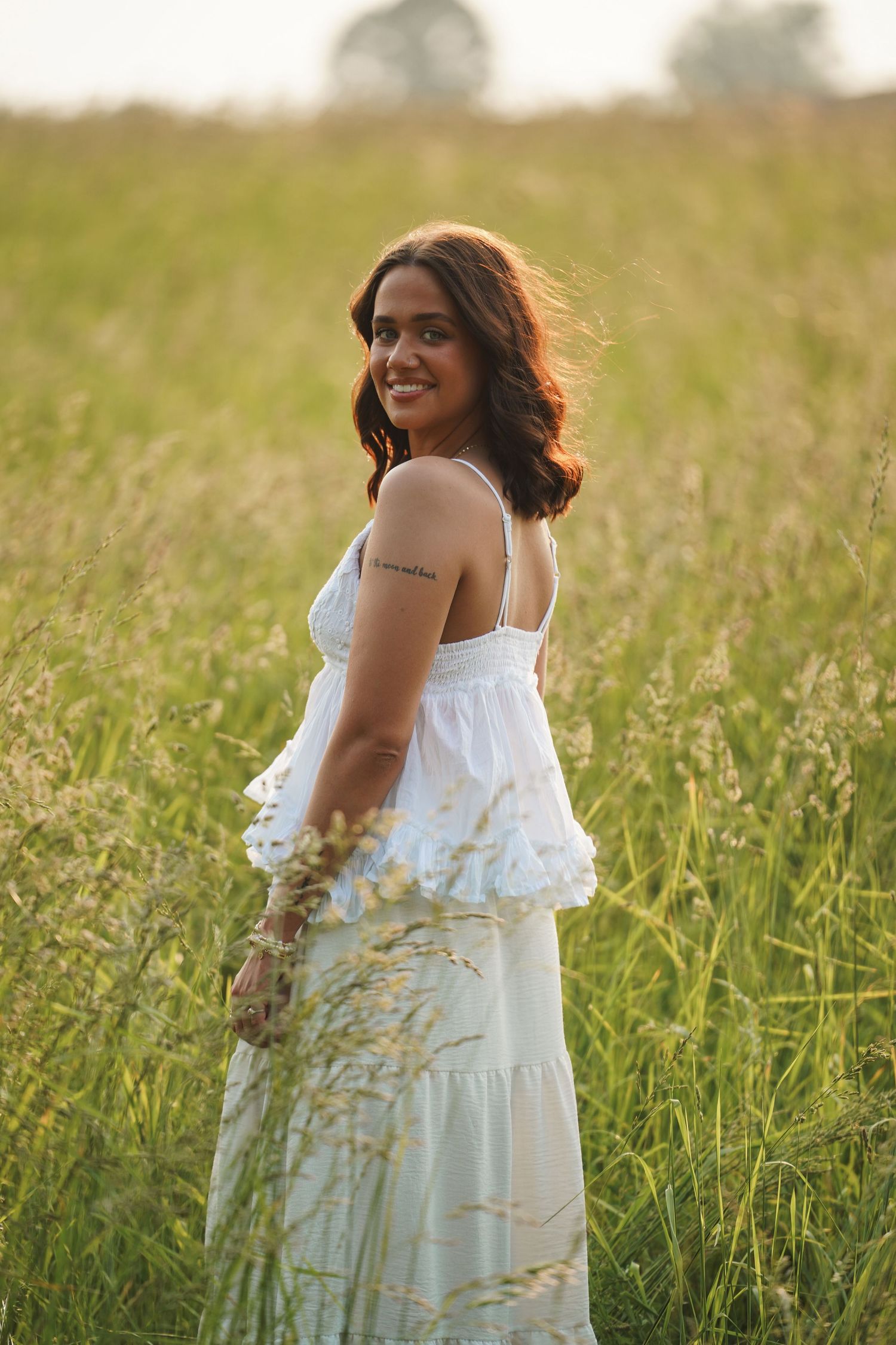 A woman wearing a white sundress smiles while standing in a golden meadow during sunset.