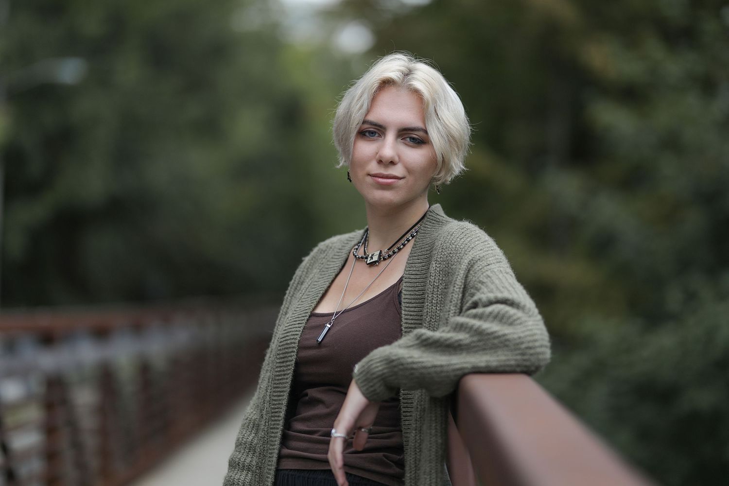A person wearing a green cardigan and brown tank top leans against a railing outdoors with greenery in the background.