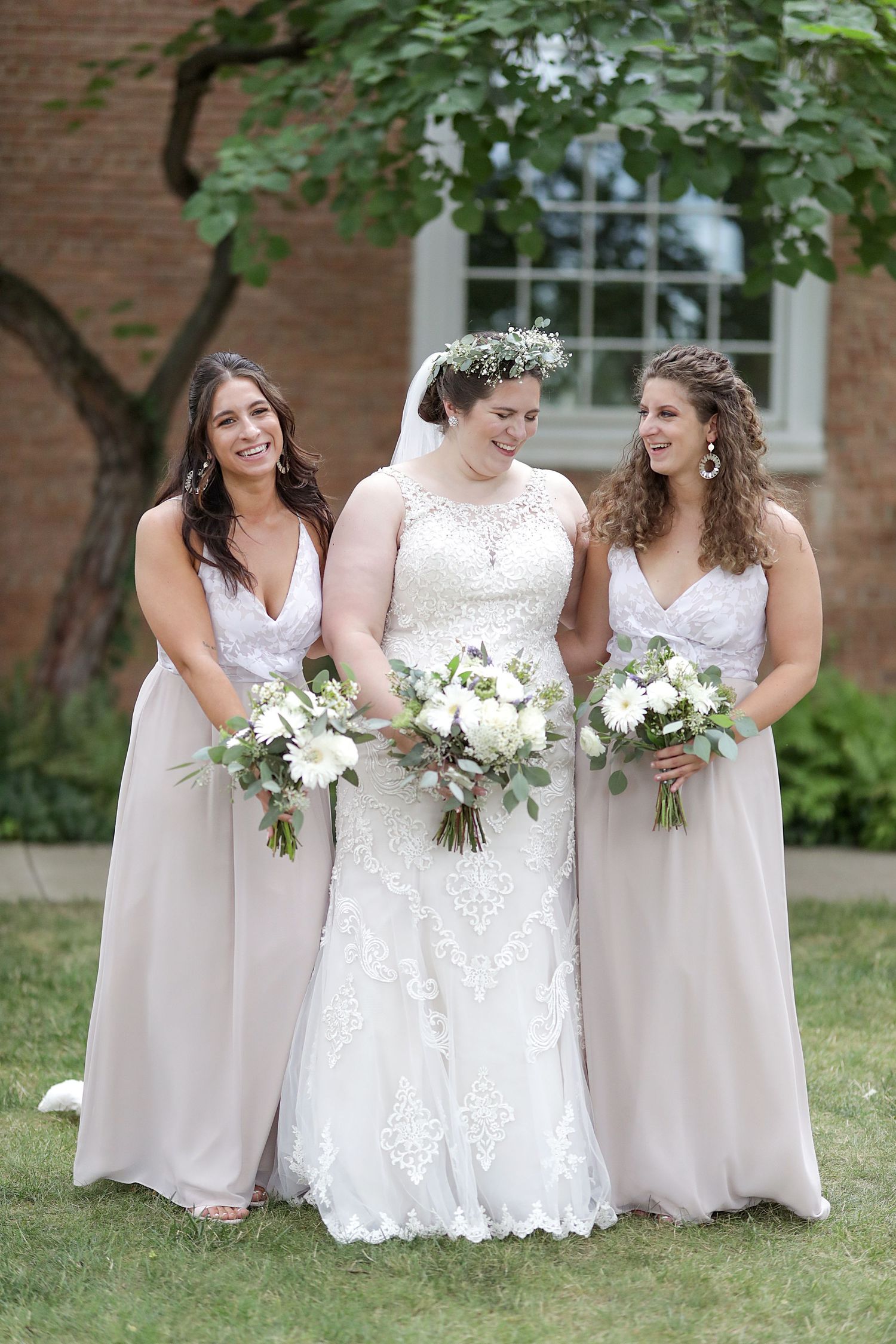 Wedding party wearing white and beige dresses share candid laughing moments while holding white floral bouquets outdoors.