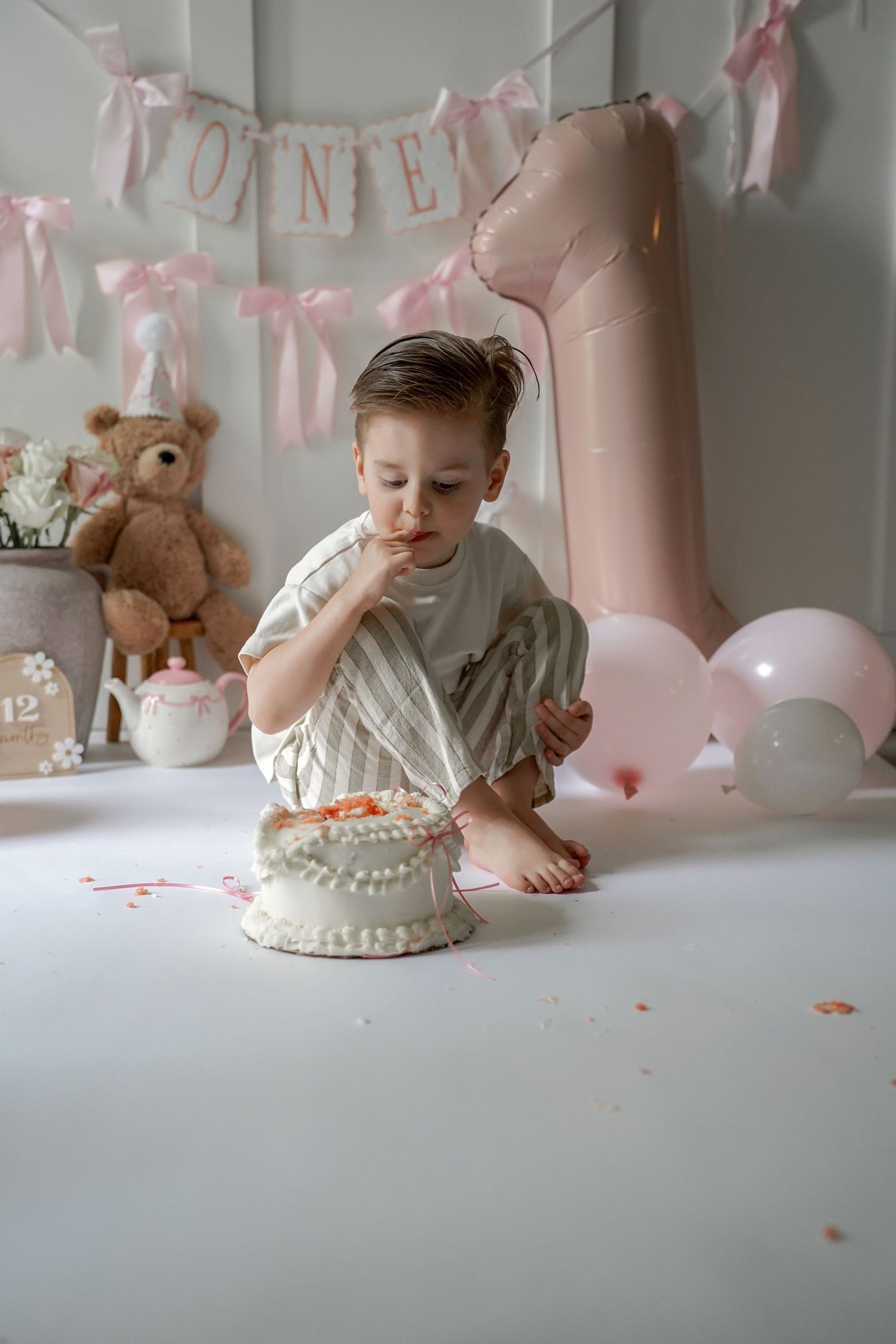 A baby's first birthday celebration with a white cake, pink decorations, and teddy bear against a soft pink backdrop.