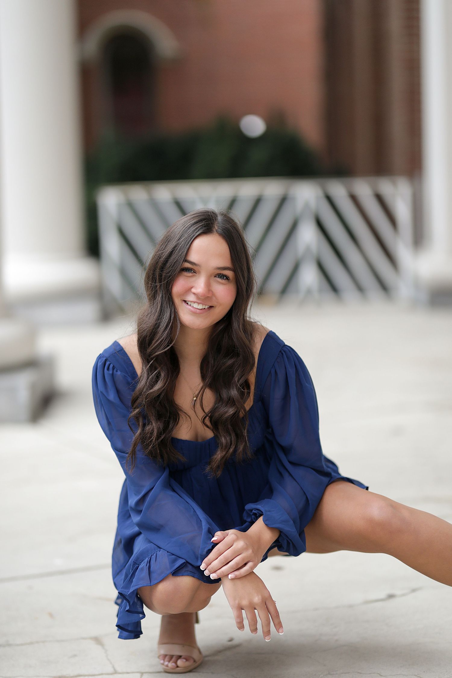 A person wearing a dark blue dress poses in a crouching position on a concrete porch with architectural columns.