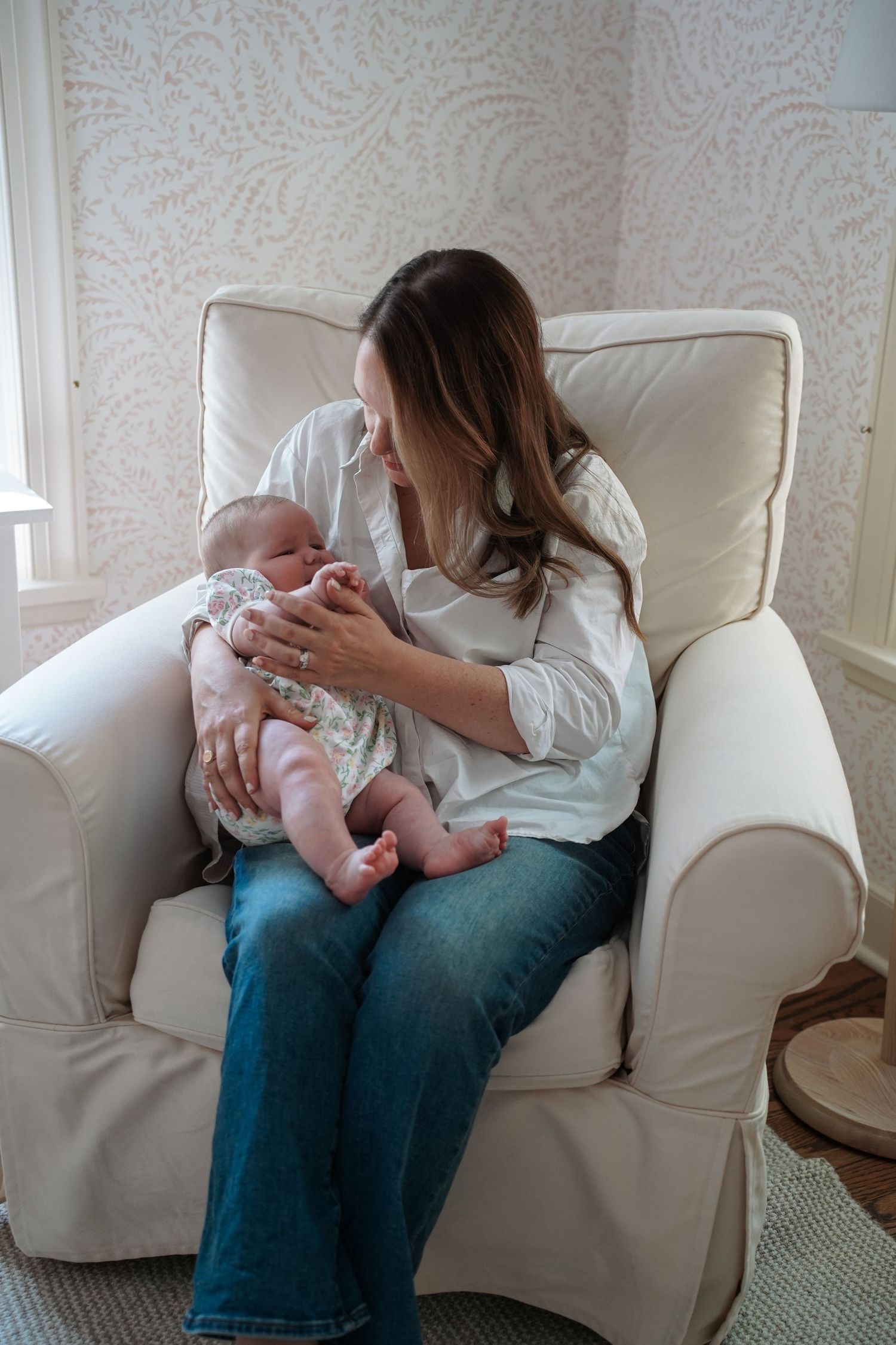 Mother and infant share a tender moment in a white armchair against patterned wallpaper.