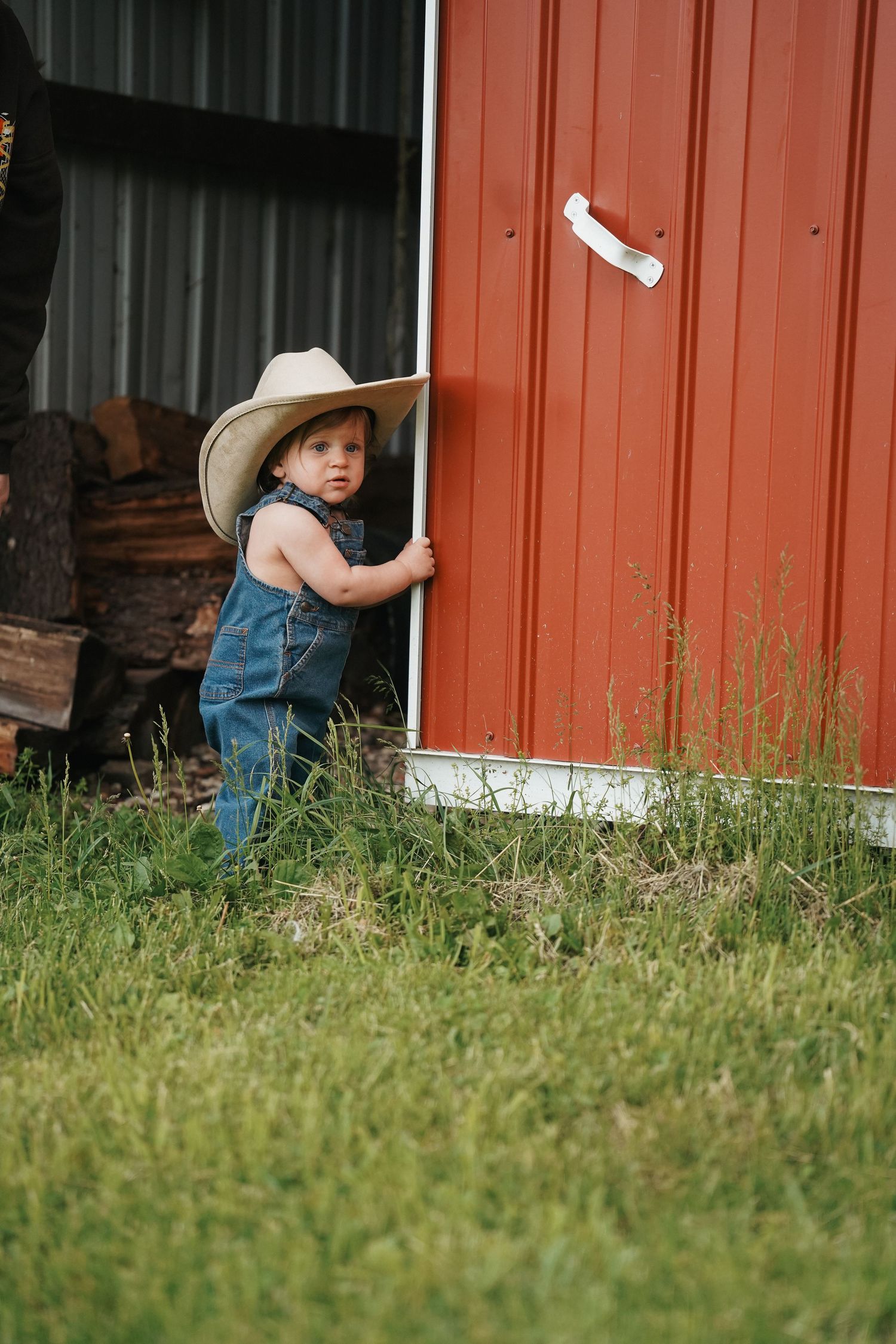 Young child wearing denim overalls and cowboy hat stands next to red barn door in grassy field.