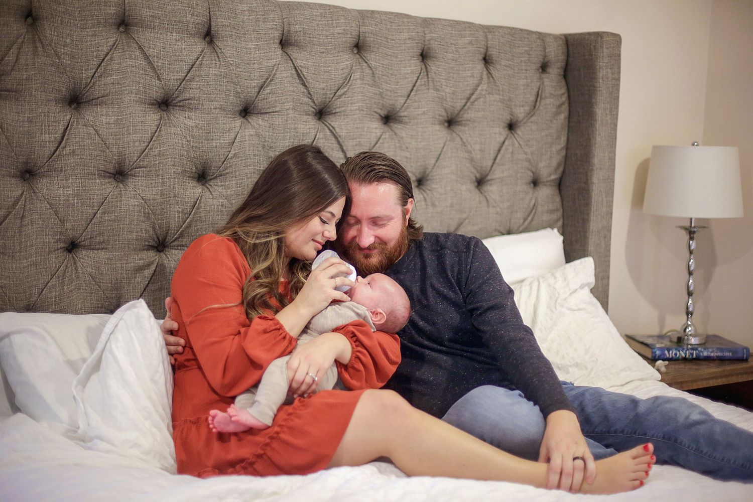 Parents snuggling with newborn baby on tufted gray headboard bed wearing cozy sweaters. Newborn photographer Chagrin Falls, Ohio.