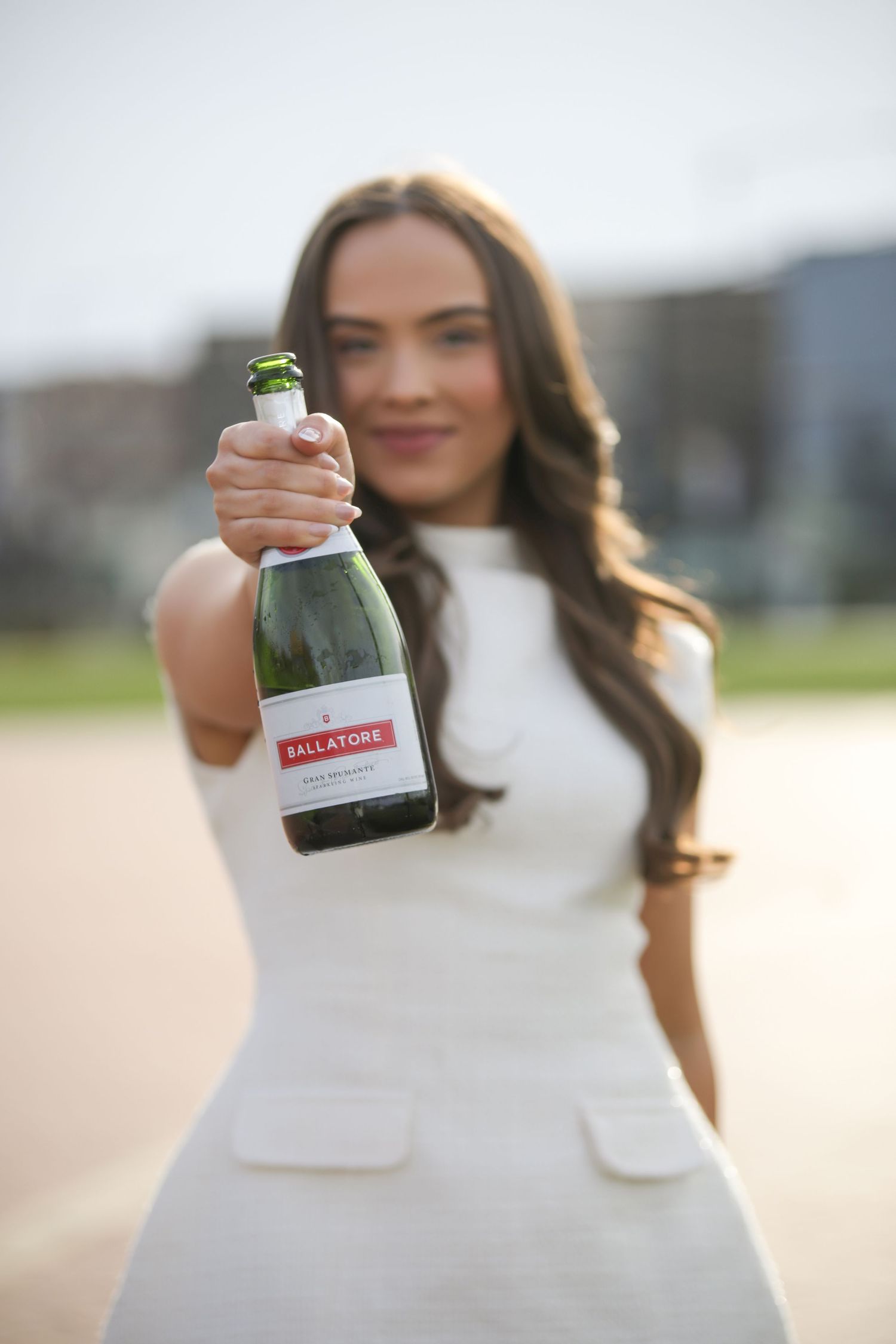 Woman in white dress holds up a small glass bottle against a blurred outdoor background.