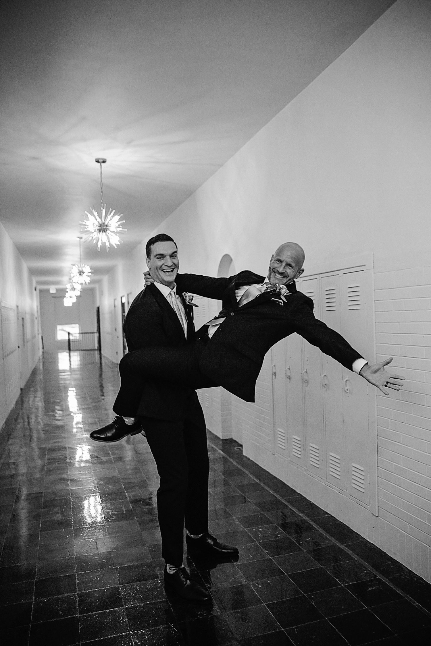 Two men in suits playfully pose in a hallway with lockers, one holding the other in a dramatic dance-like position.