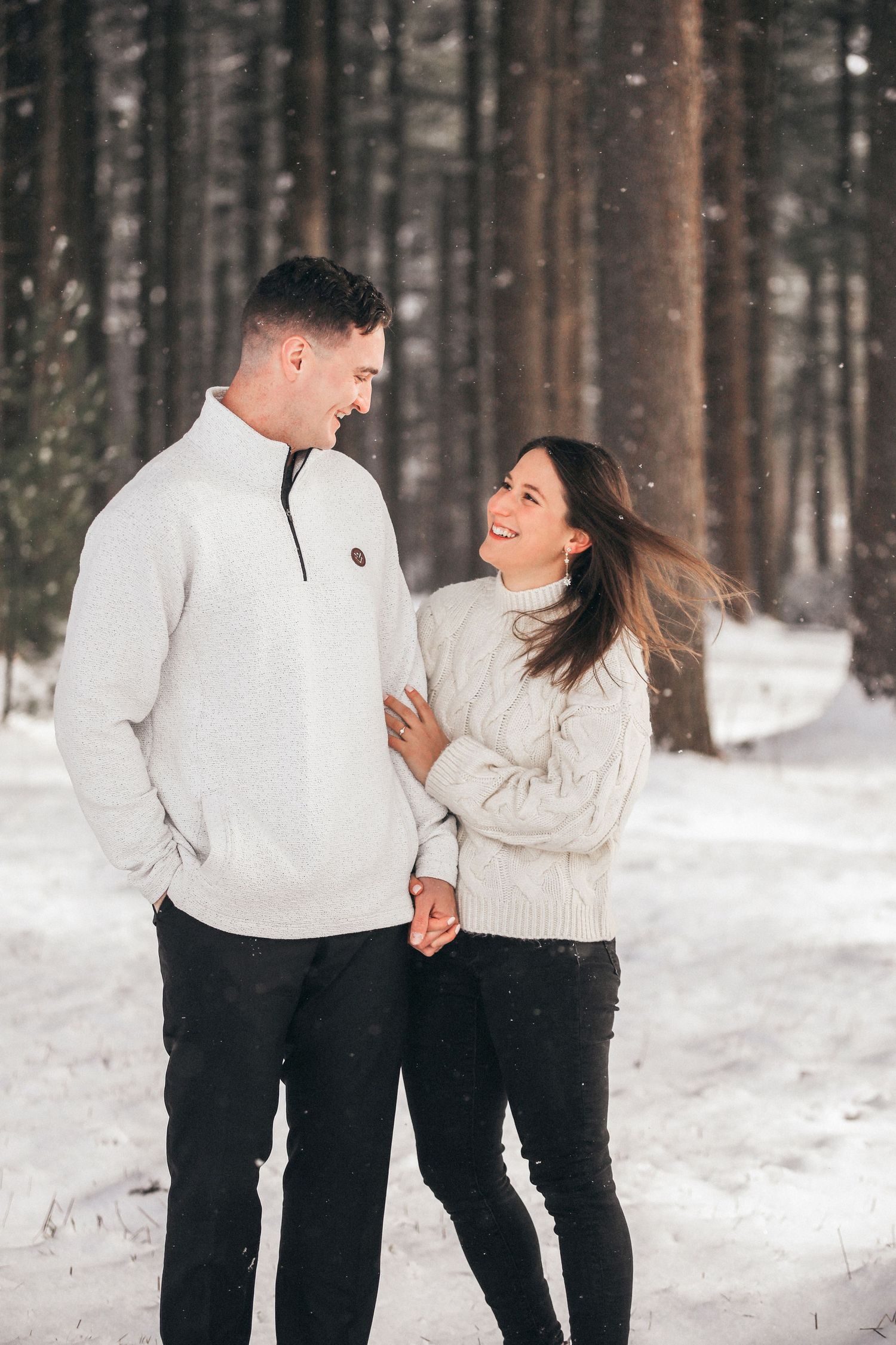 A couple in matching white sweaters and black pants share playful moments together in a snowy winter forest setting.