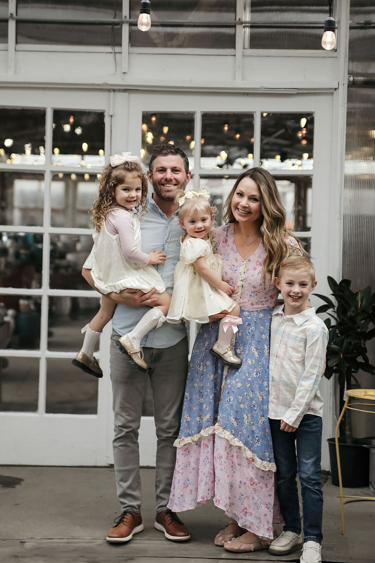 A family poses together in a casual setting with glass windows and string lights in the background.