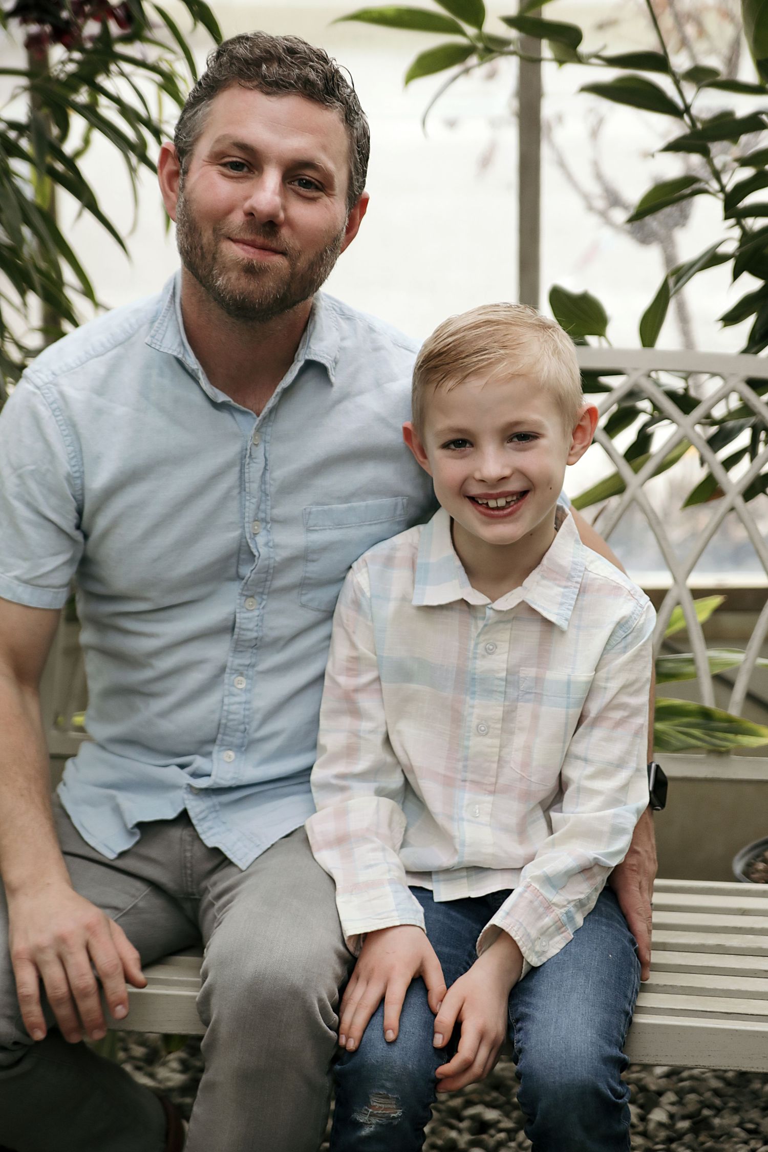 A parent and child sit together inside a greenhouse with tropical plants in the background.
