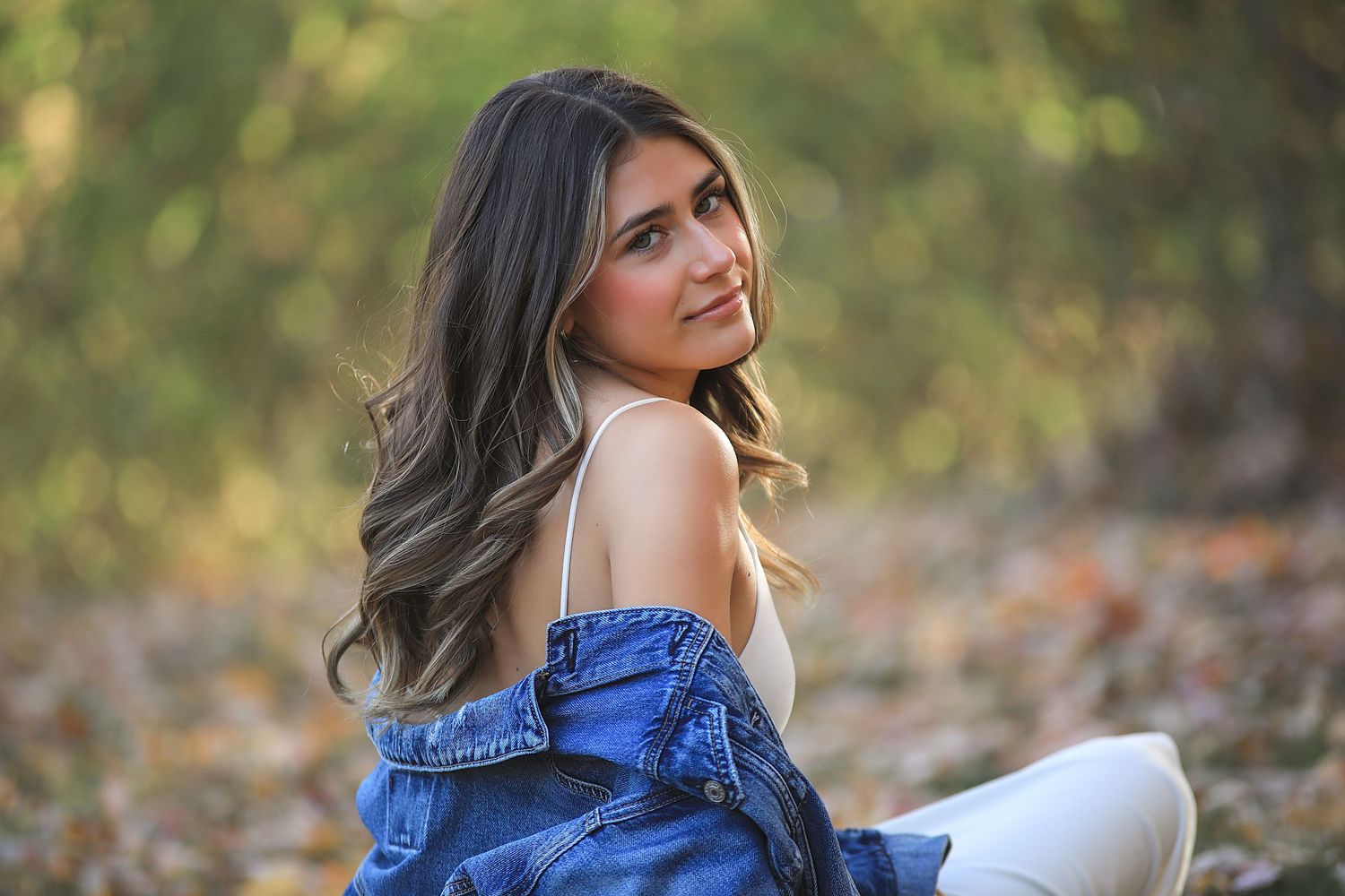 A smiling woman in a blue denim top poses outdoors in autumn with trees and fallen leaves in the background.
