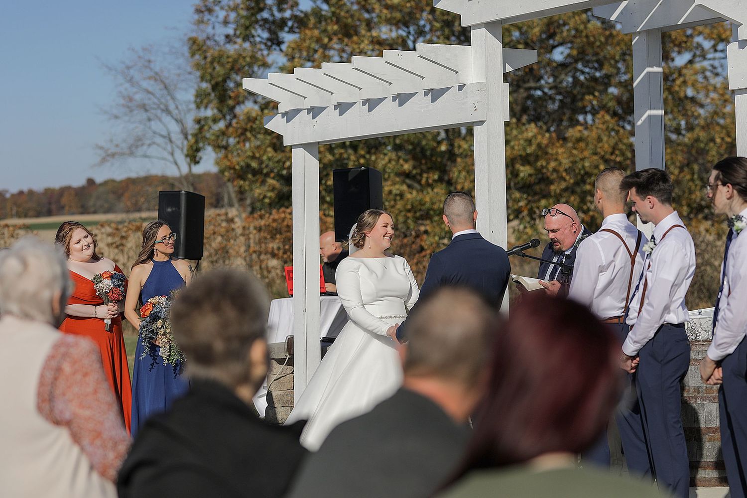 Outdoor wedding ceremony taking place under white pergola with fall foliage in background.