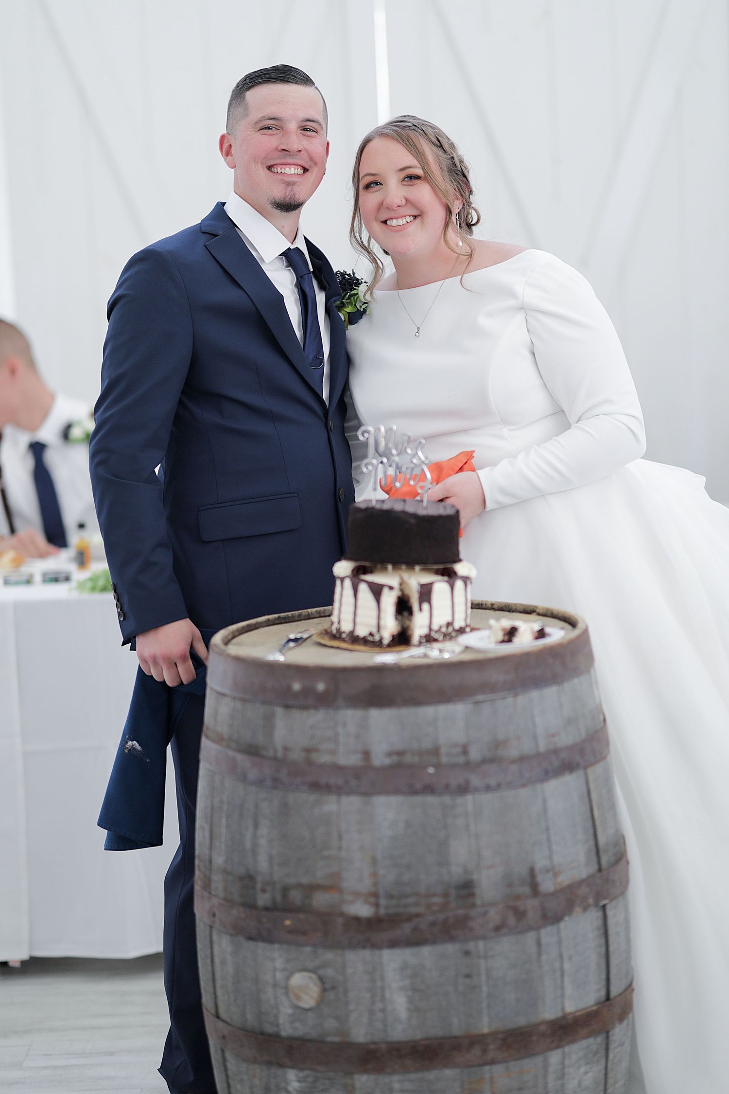 Wedding couple poses with cake on rustic wooden barrel at indoor reception venue.