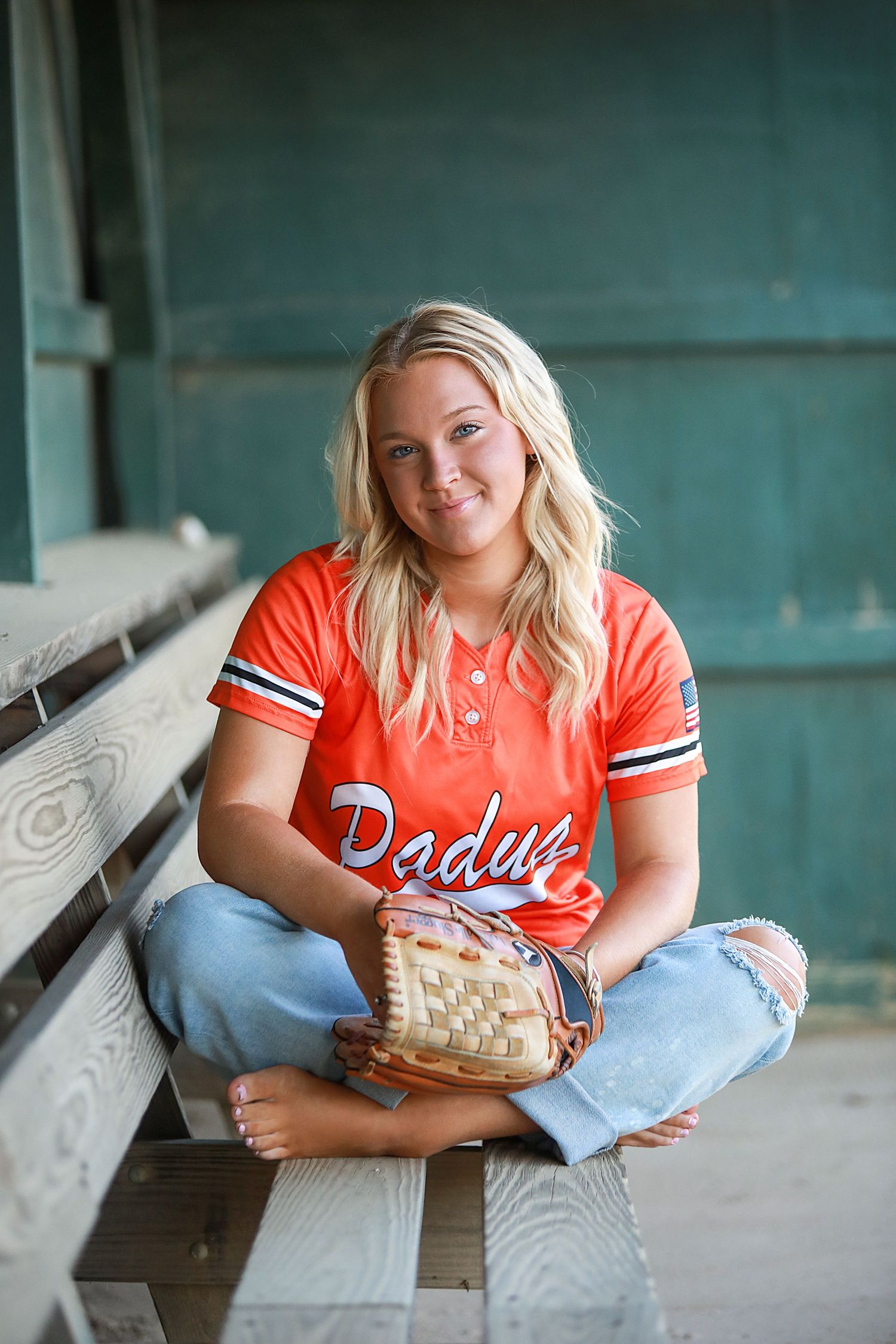 A person in a red baseball jersey and ripped jeans sits in a dugout holding a baseball glove. Senior portraits Brecksville, Ohio