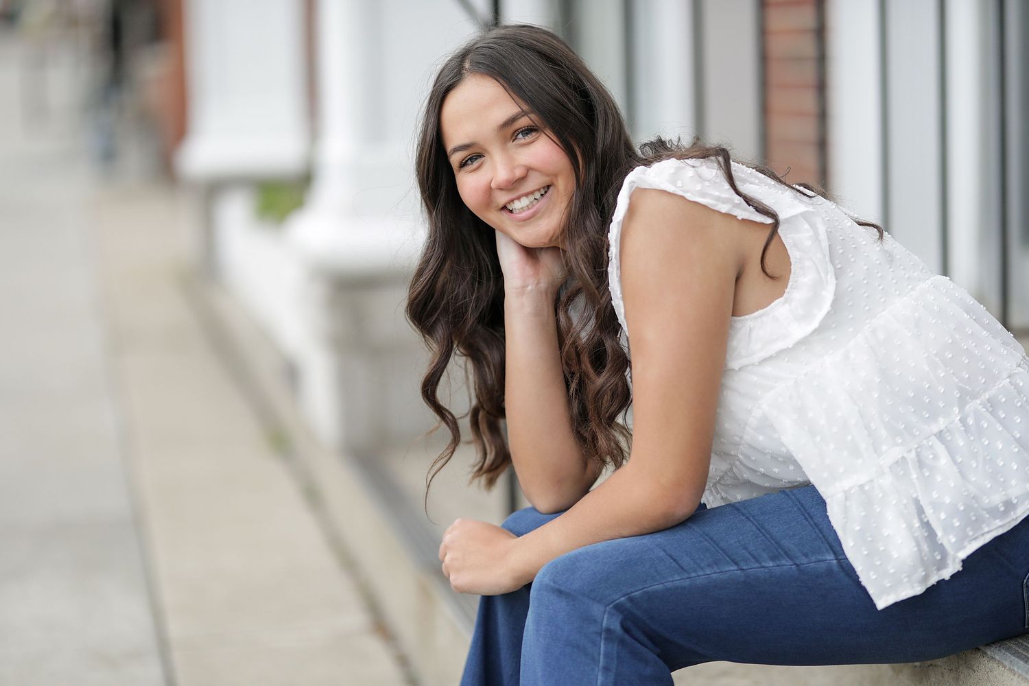 Young woman in white sleeveless top and blue jeans sits casually on outdoor steps and smiles at camera.