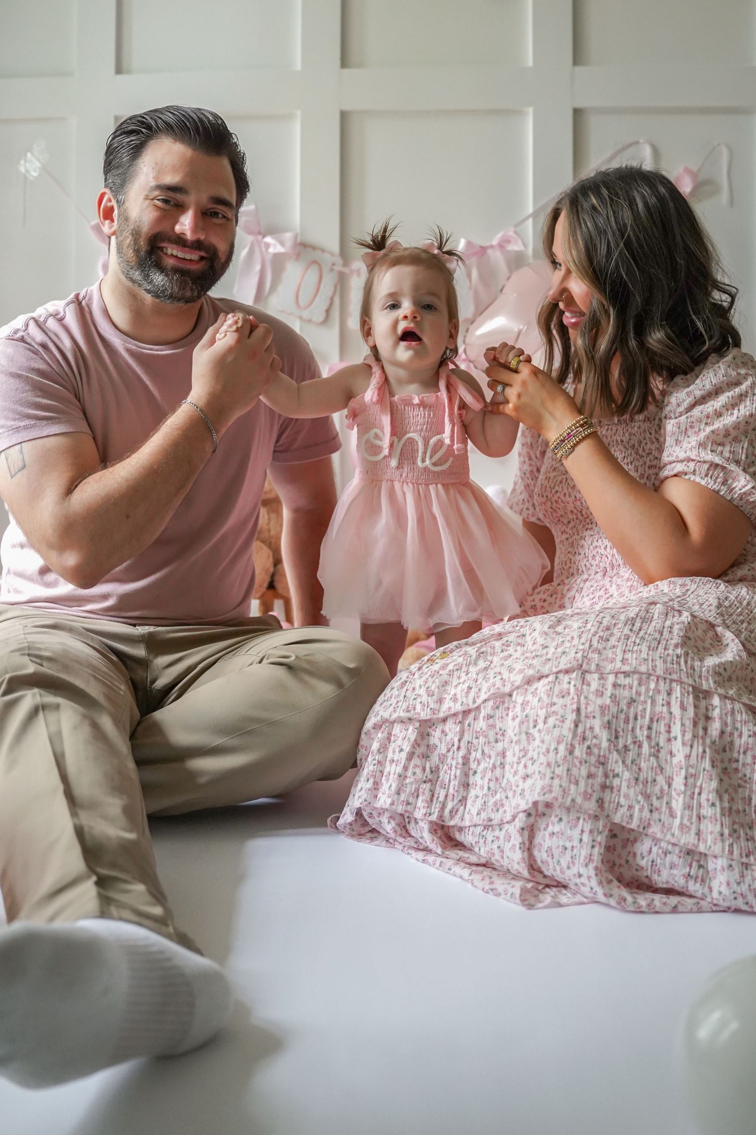 A family sits together on a couch wearing pink and neutral tones while sharing a joyful moment.