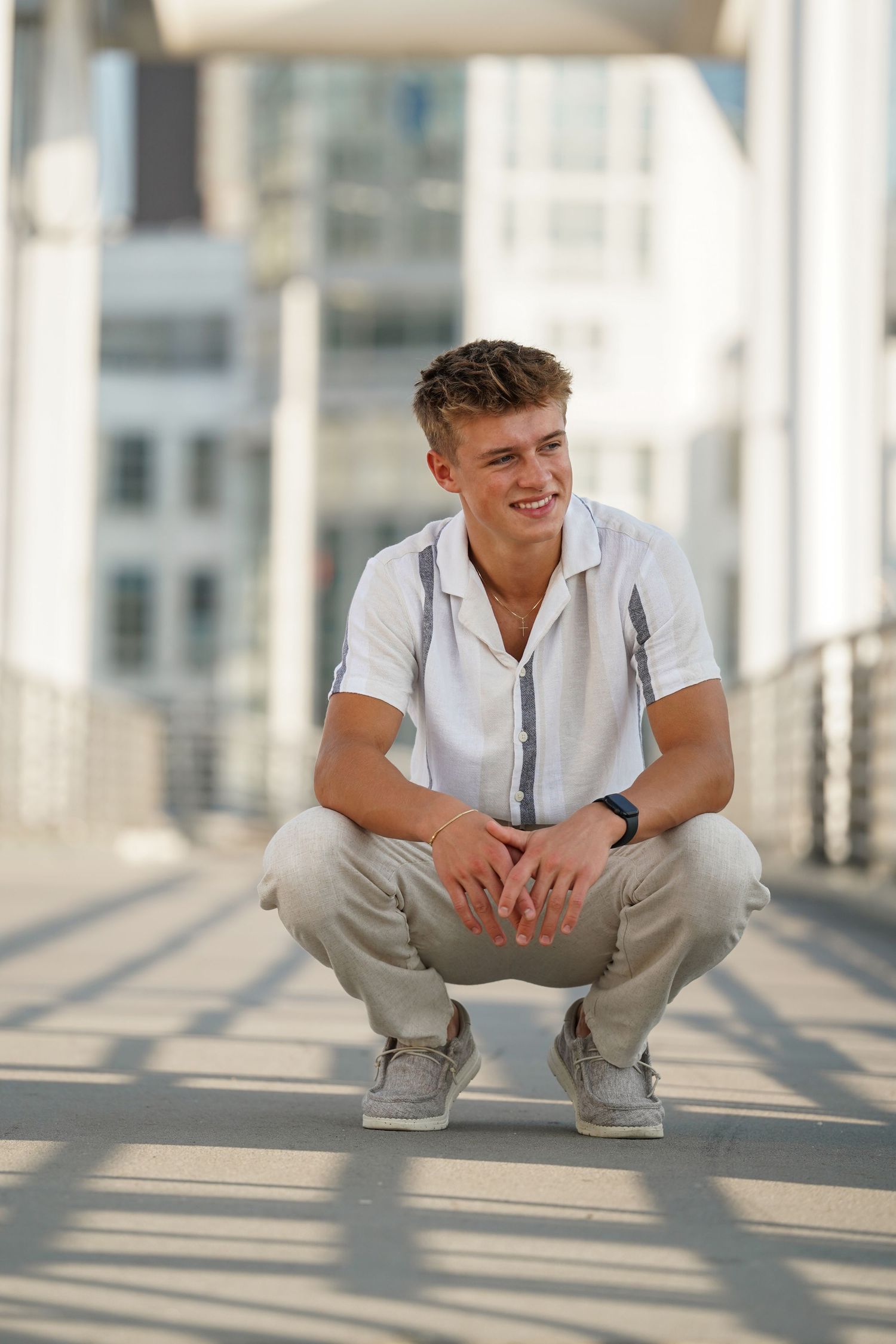 Portrait of a young person in a white polo shirt and khaki pants crouching on a bright walkway with urban architecture behind.
