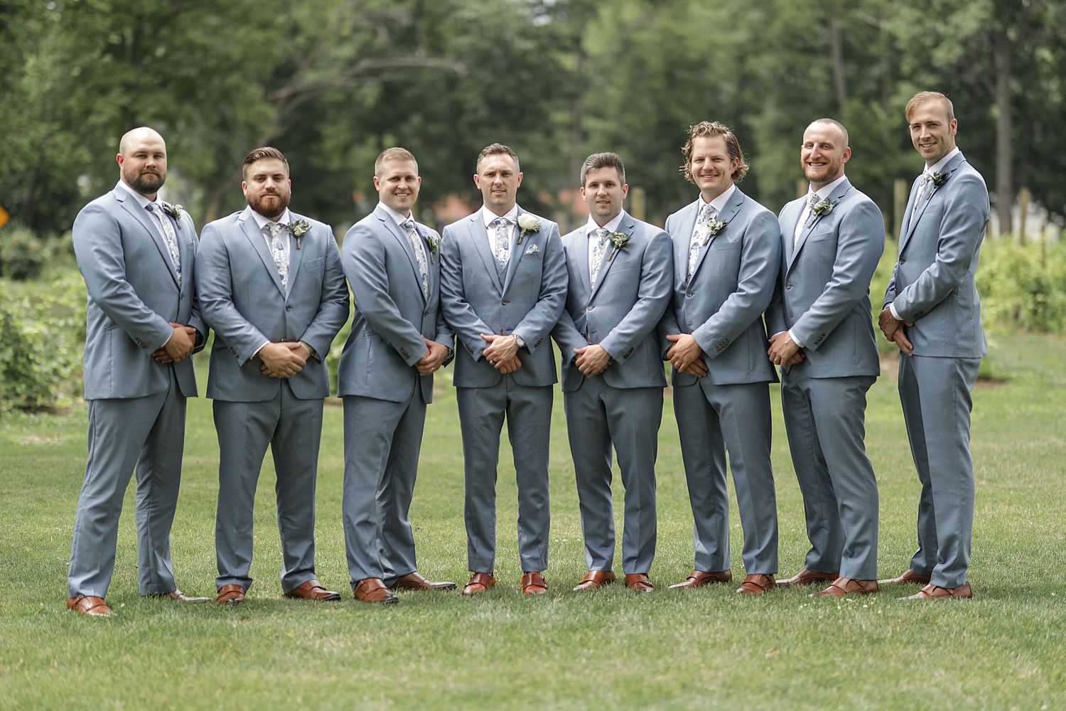 Groomsmen in gray suits pose playfully on grass in a line formation.