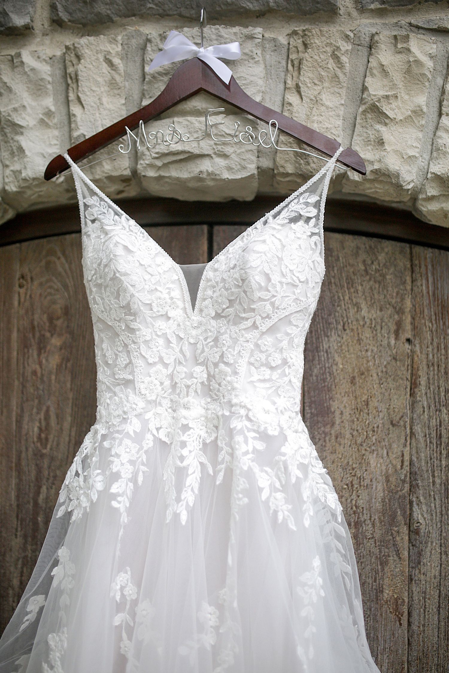 Elegant white lace wedding dress with plunging neckline displayed on wooden hanger against stone wall.