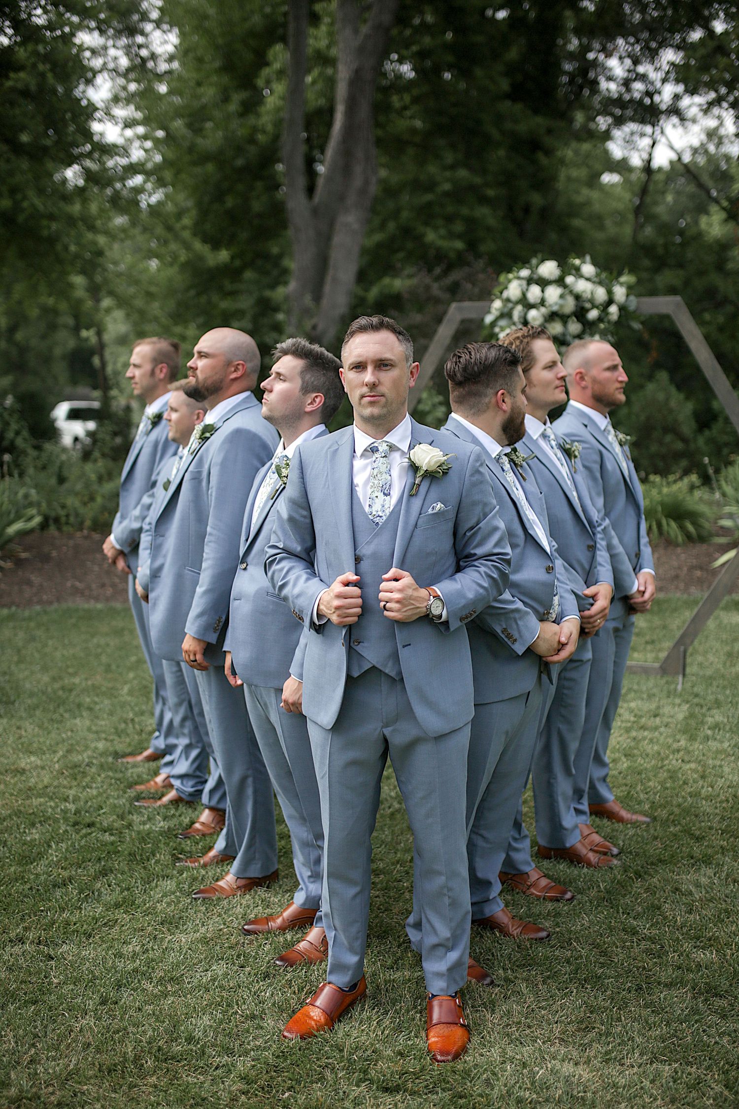 Wedding party in coordinated grey suits and brown shoes pose in V-formation on green lawn.