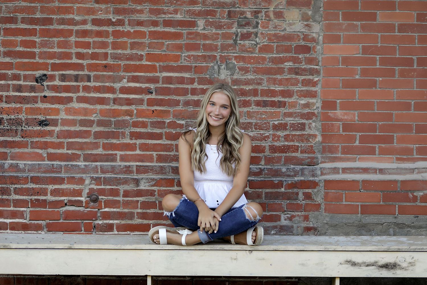 Young woman in white top and jeans sits cross-legged against rustic brick wall background.