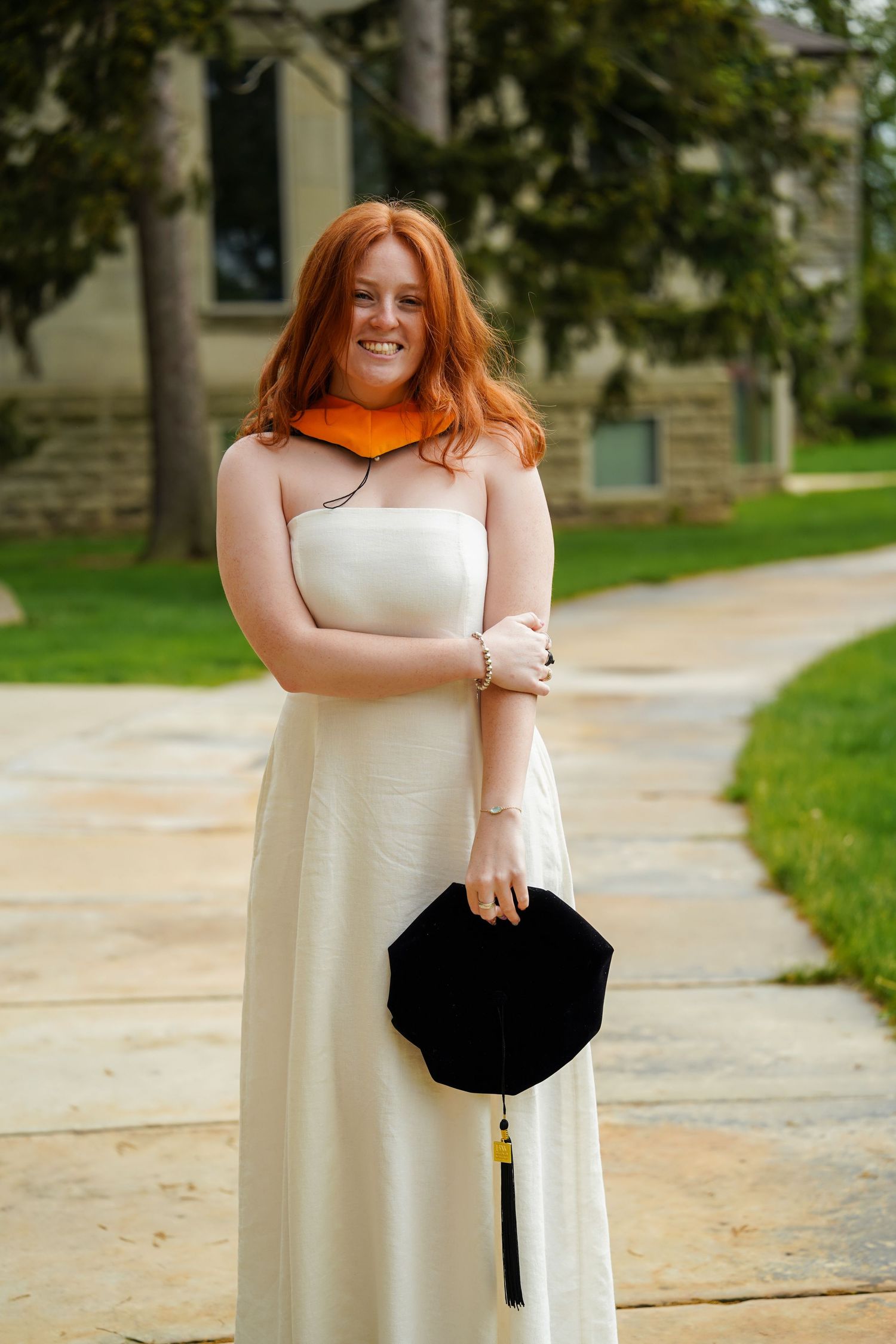 Person in a flowing white formal dress and orange neckpiece holding a black clutch purse stands on a stone walkway.