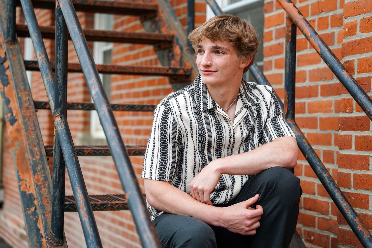 Someone sits casually on metal stairs against a brick wall wearing a striped button-down shirt and dark pants.