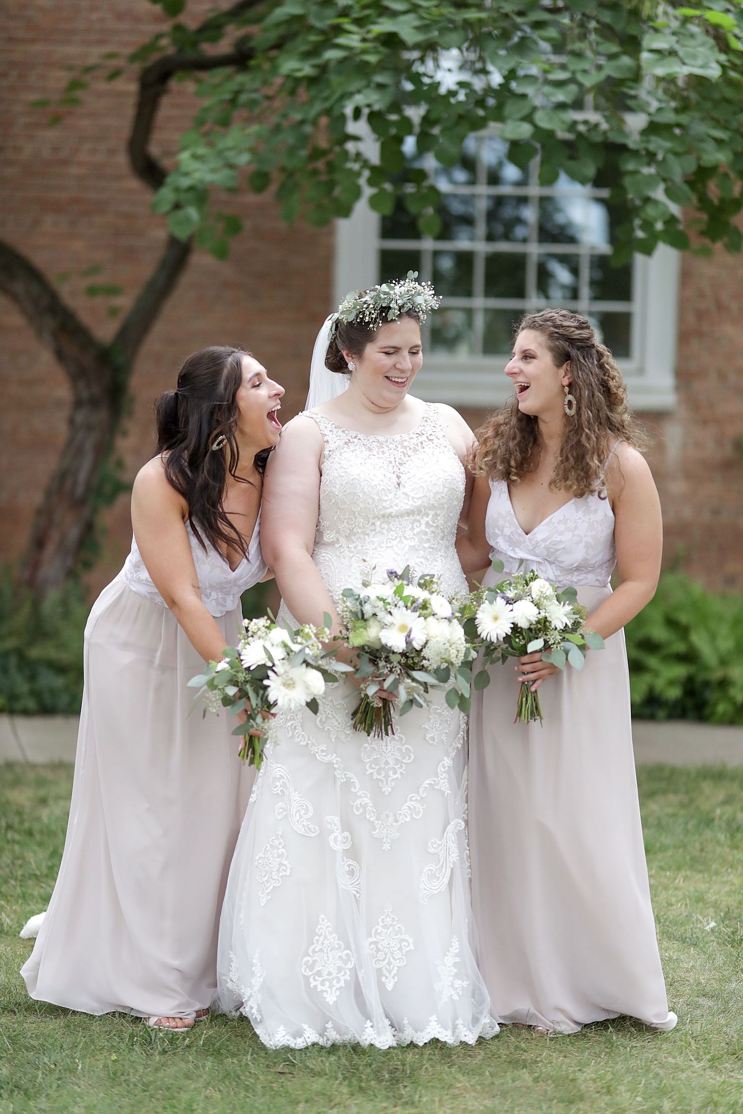 A group in pale pink bridesmaid dresses and white wedding gown share joyful laughter outdoors at a garden ceremony.
