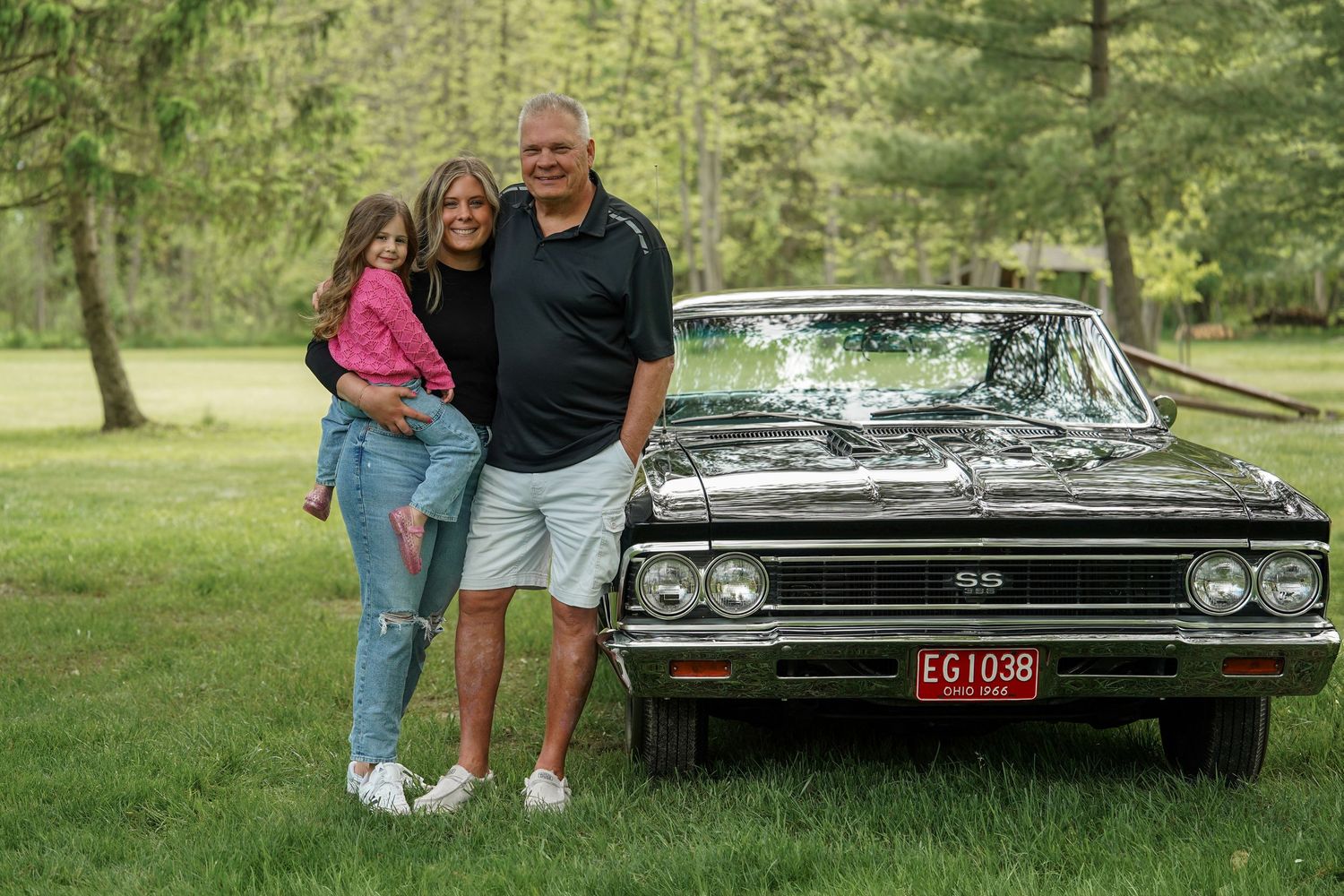 Two people stand beside a black vintage car in a park setting.