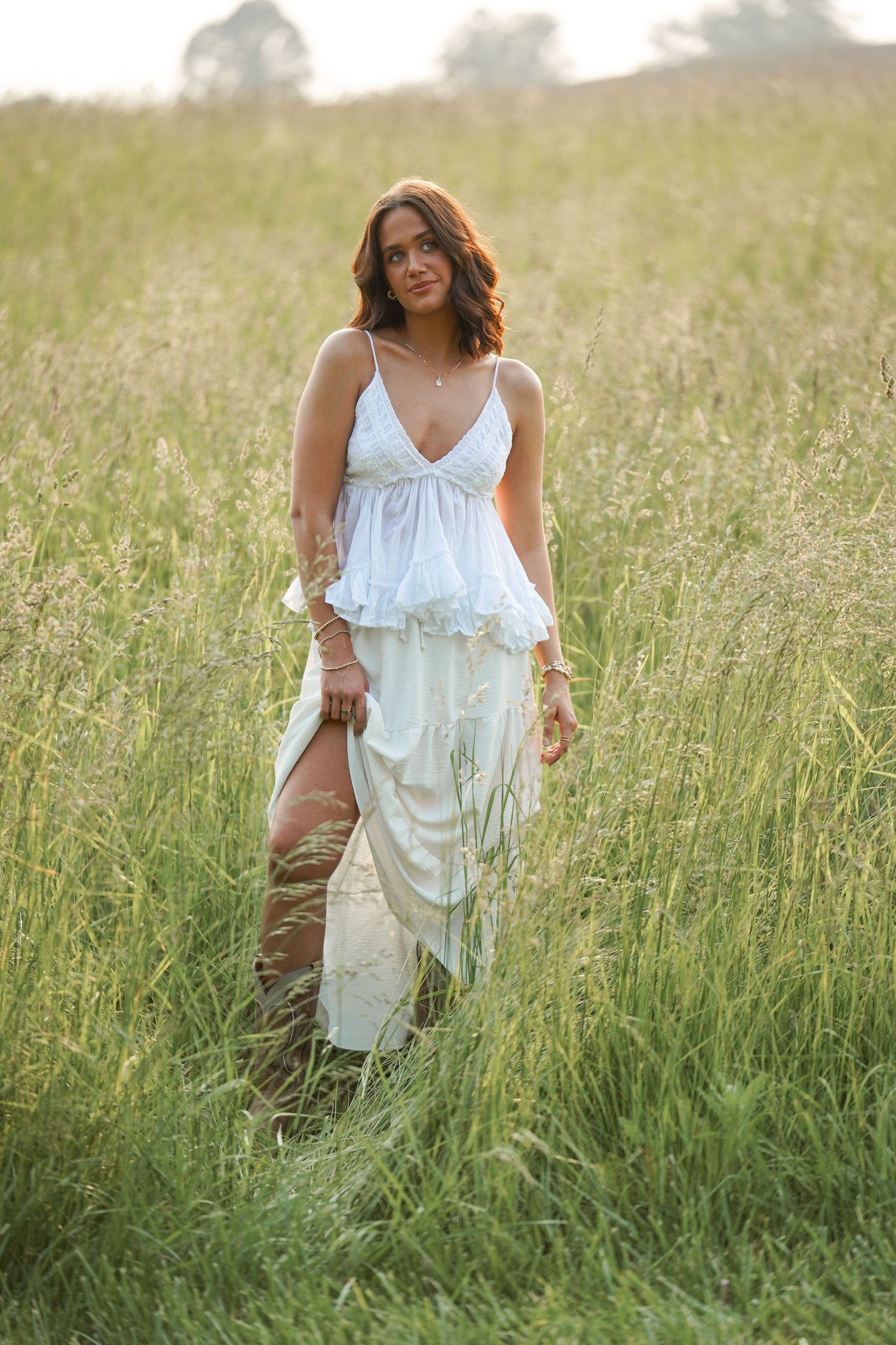 A woman in a flowing white summer dress walks through a sunlit field of tall grass at golden hour.