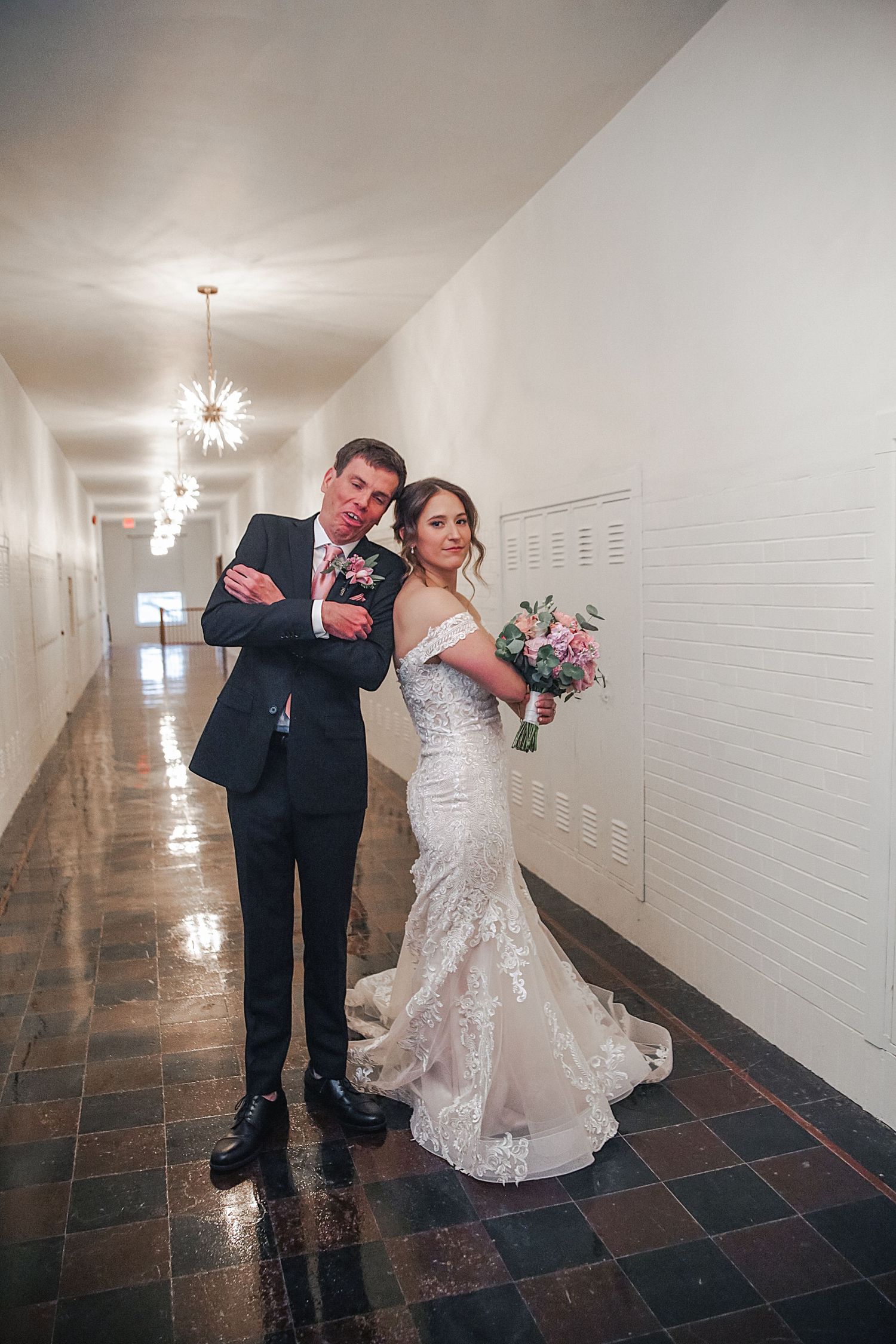 A bride and groom share a playful moment in a white-walled hallway with tiled floors.