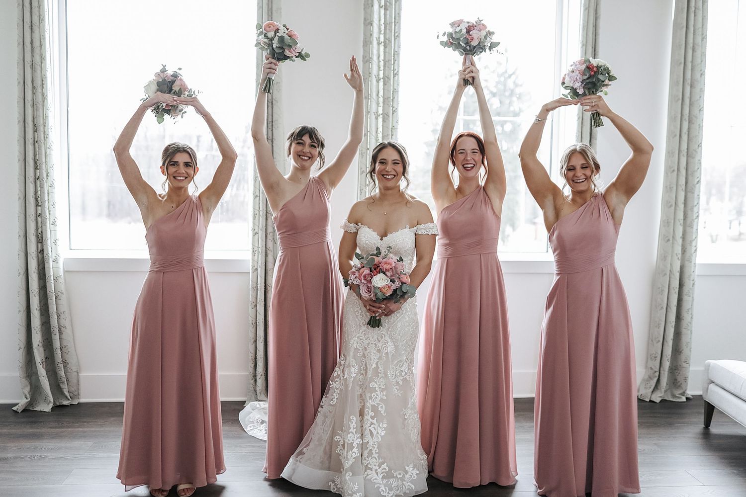 Bridesmaids in dusty rose dresses pose together holding bouquets in an elegant indoor setting.
