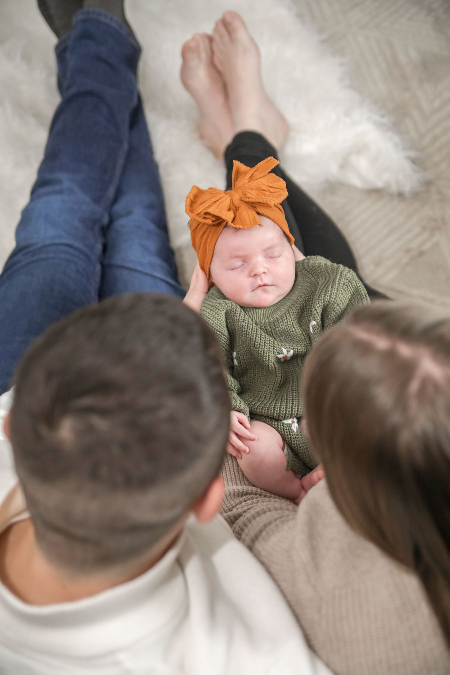 Baby wearing green sweater and orange headband lies on white blanket while being watched from above.
