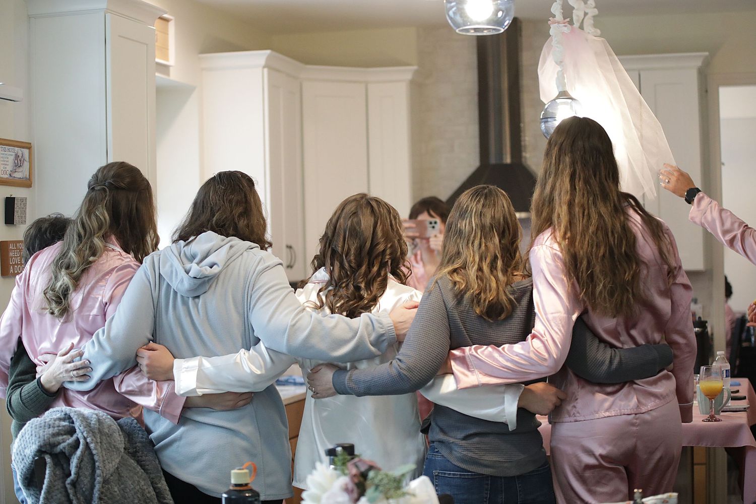 A group of people huddle together in a kitchen, wearing casual loungewear in soft pastel colors.