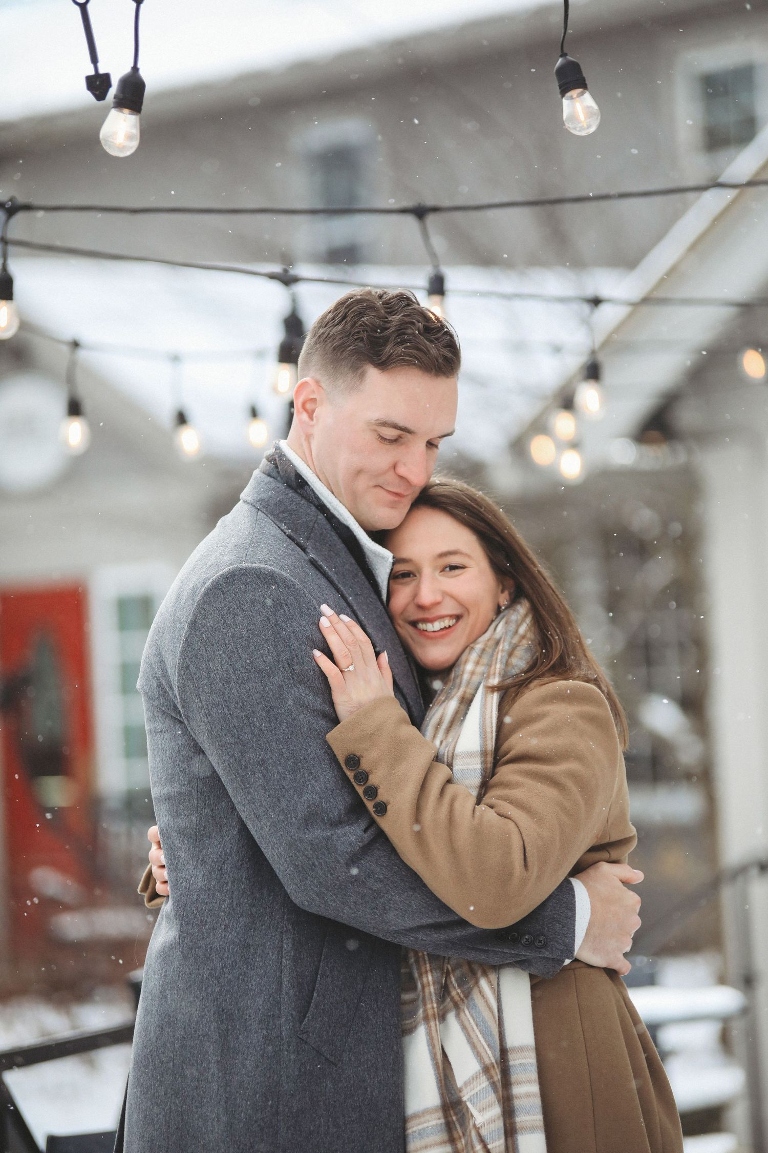 A couple shares a tender embrace outdoors in winter with string lights glowing in the background.