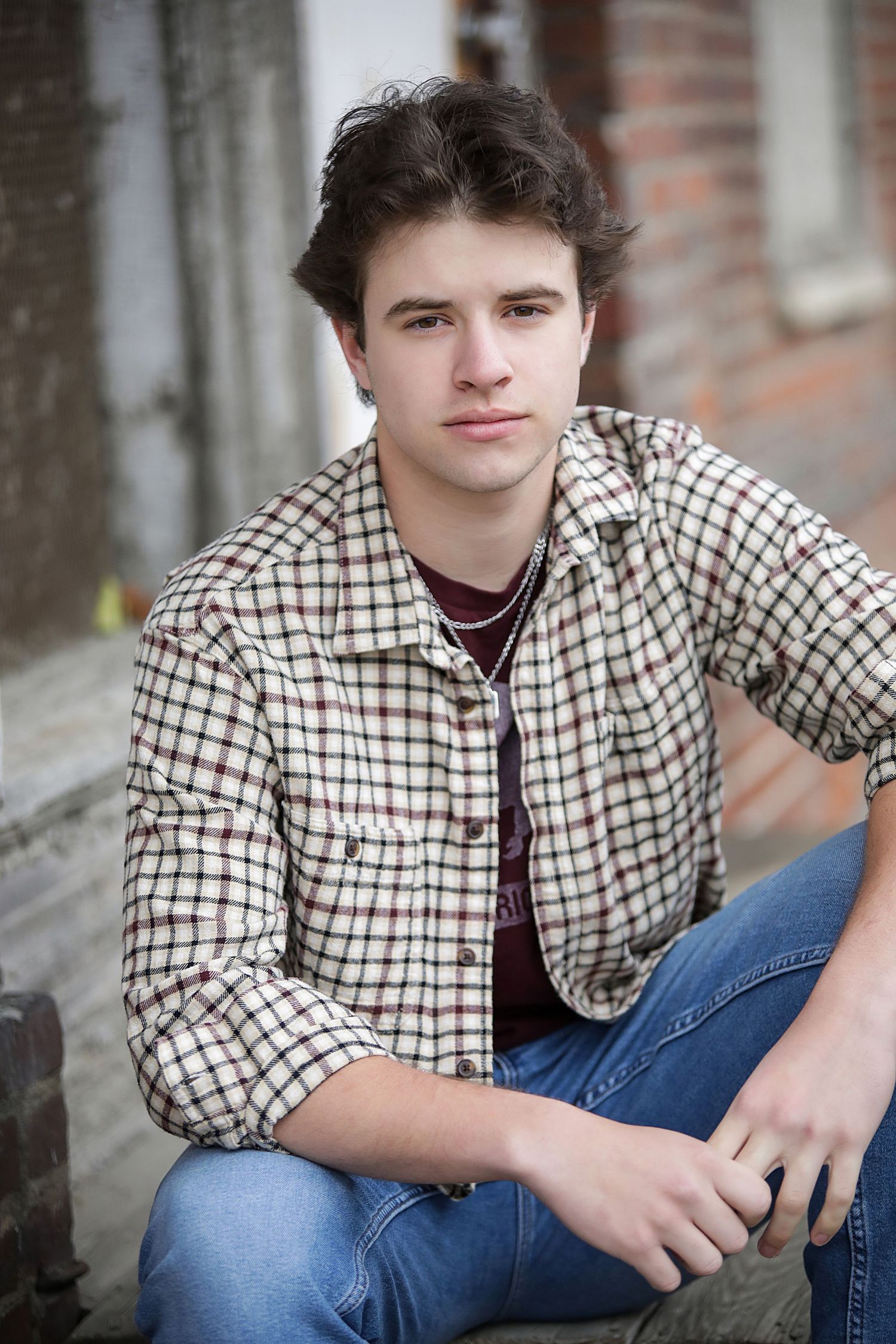 A person in a checkered button-down shirt and jeans sits casually against a brick wall background.