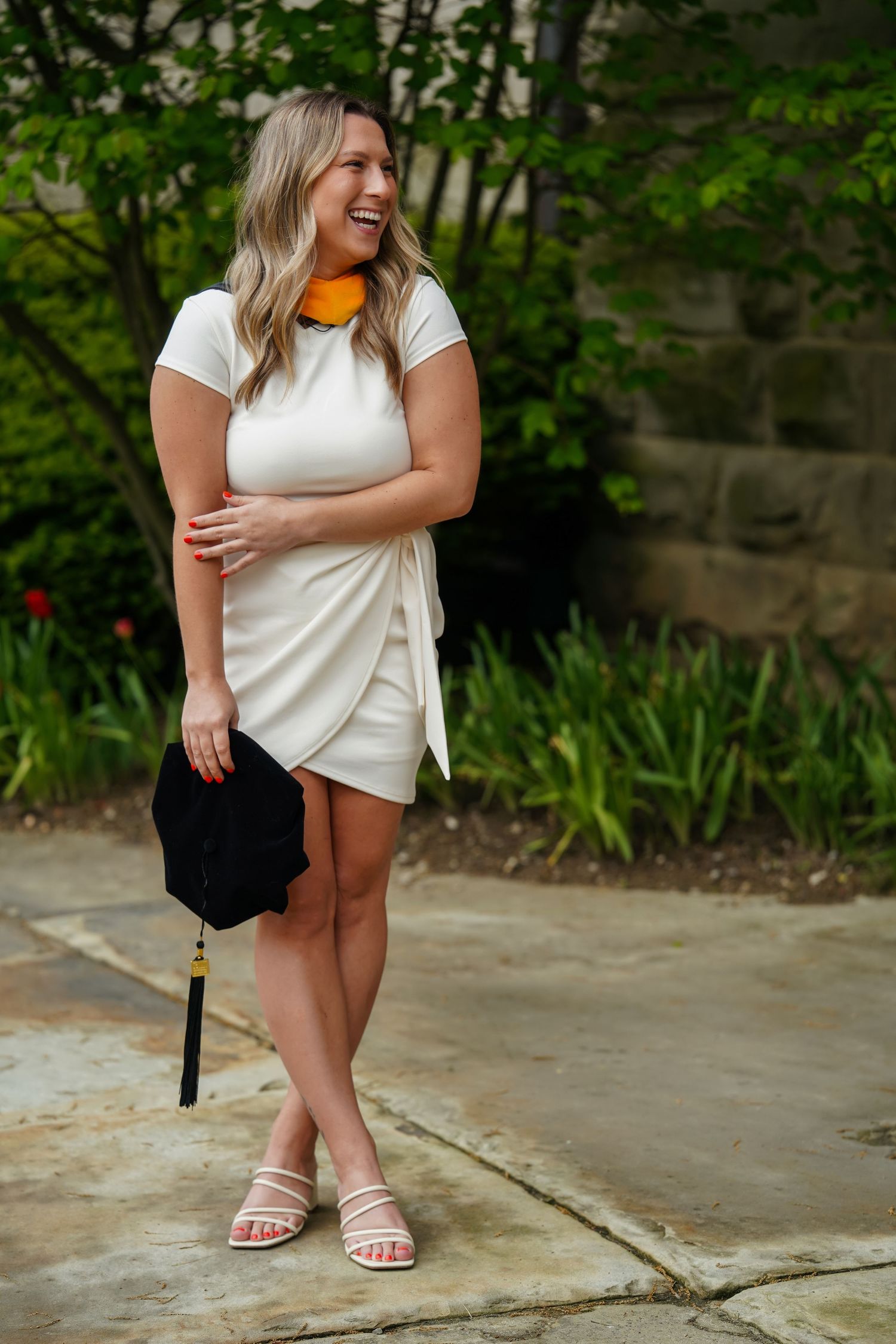 Woman in white wrap dress and orange scarf smiles while standing outdoors against stone wall and greenery.
