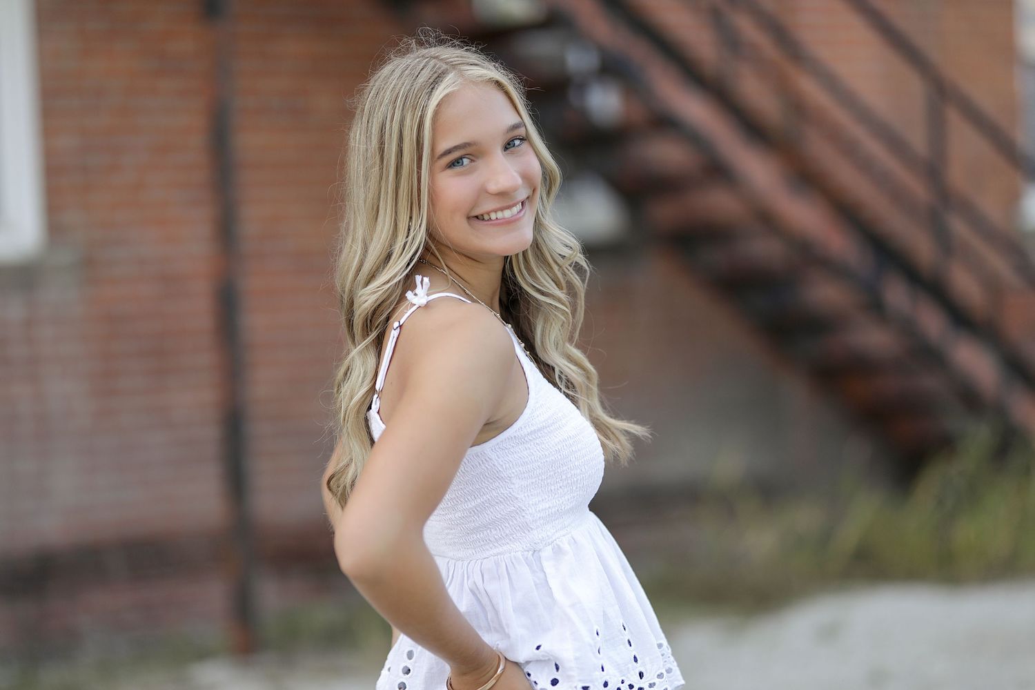 Person in white eyelet lace dress poses outdoors against brick wall with metal staircase in background.