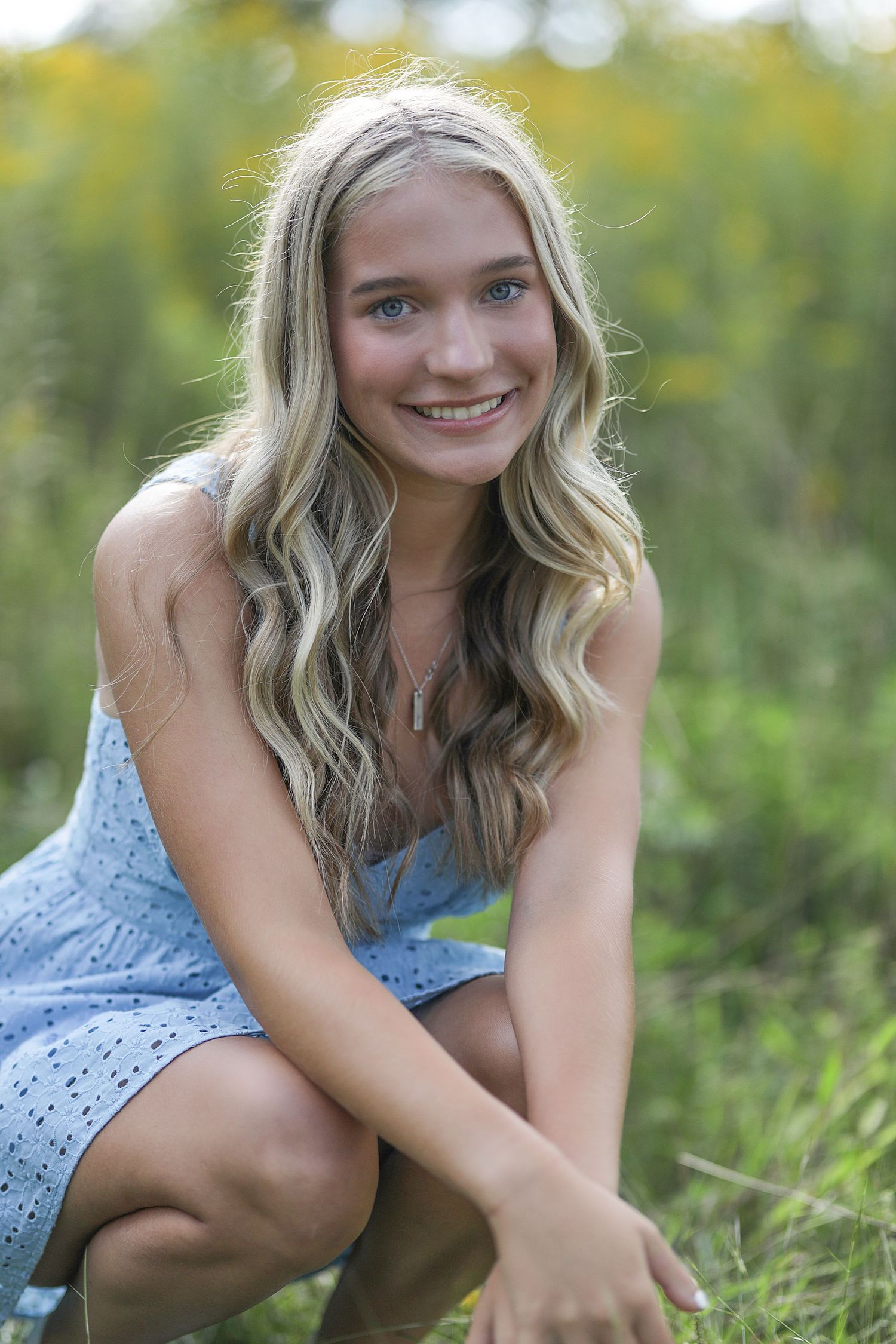 A smiling young woman wearing a light blue dress crouches in green grass with blurred natural background.
