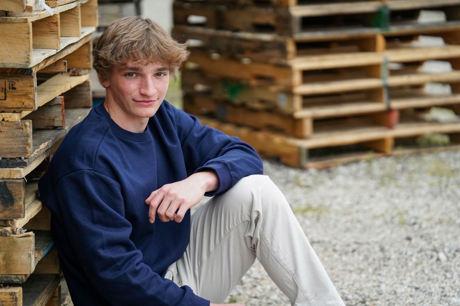 A person in a navy blue shirt sits casually on a bed of gravel with wooden pallets in the background.