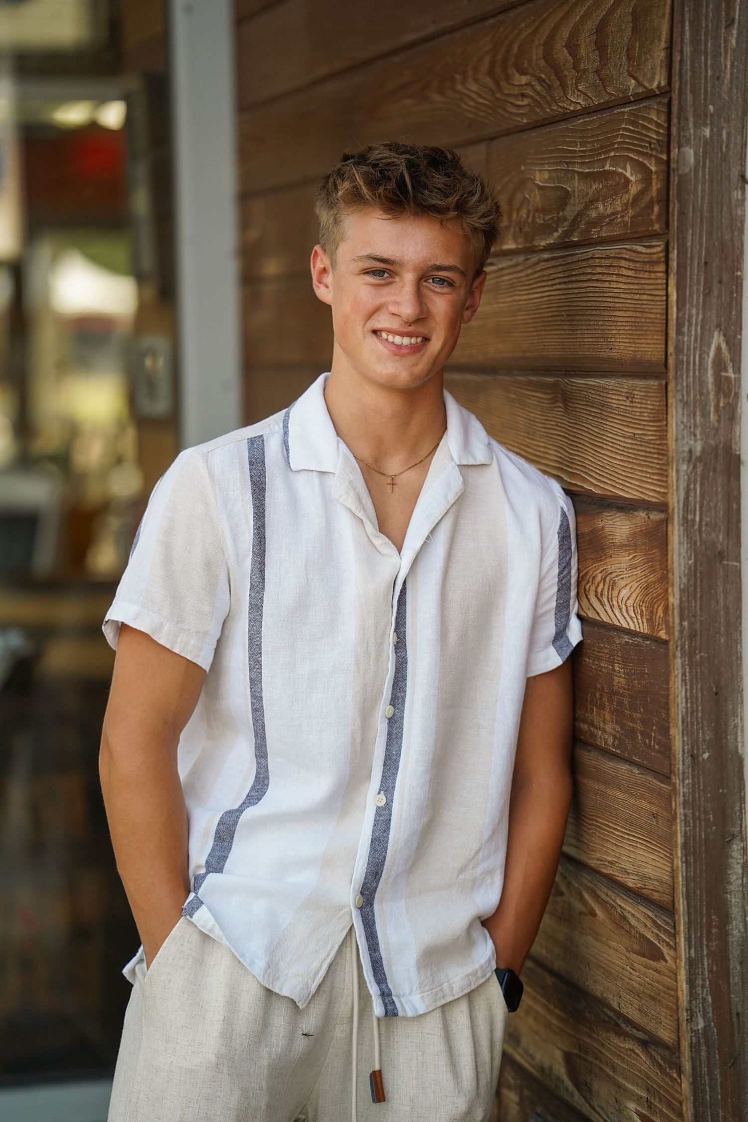 Young person wearing a white button-down shirt poses against rustic wooden wall panels.