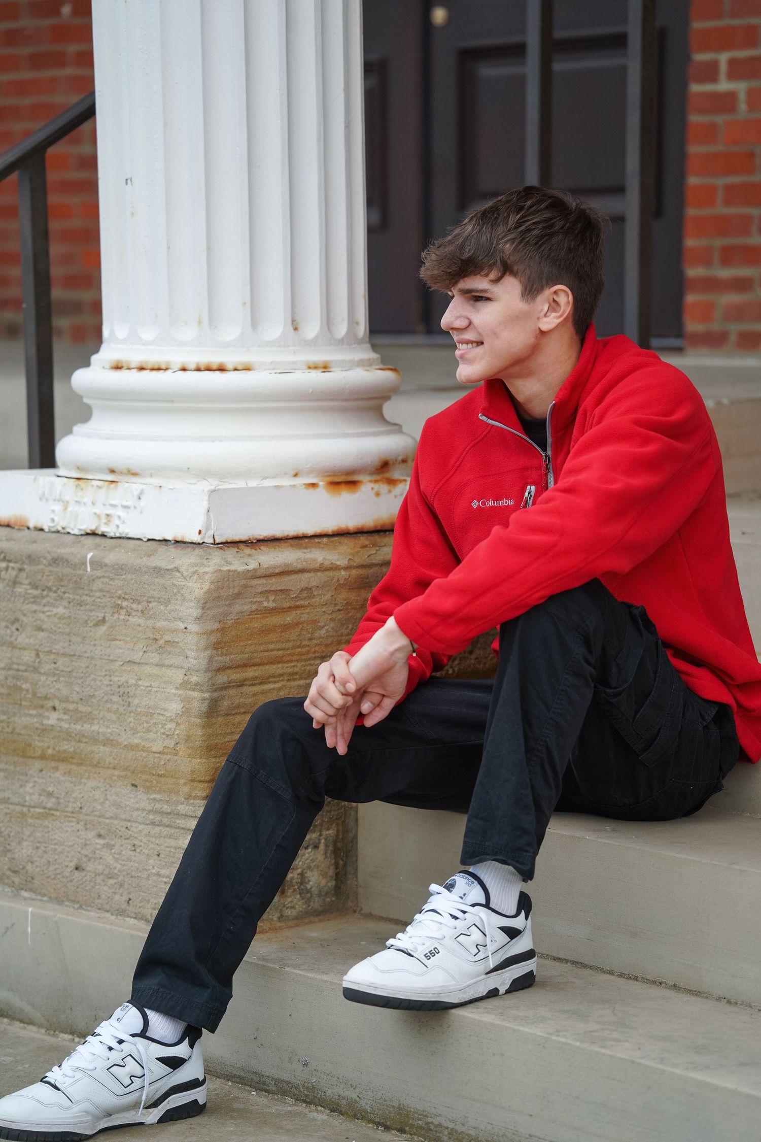 A person in a red sweatshirt and dark jeans sits on stone steps next to a white column in a casual outdoor pose.